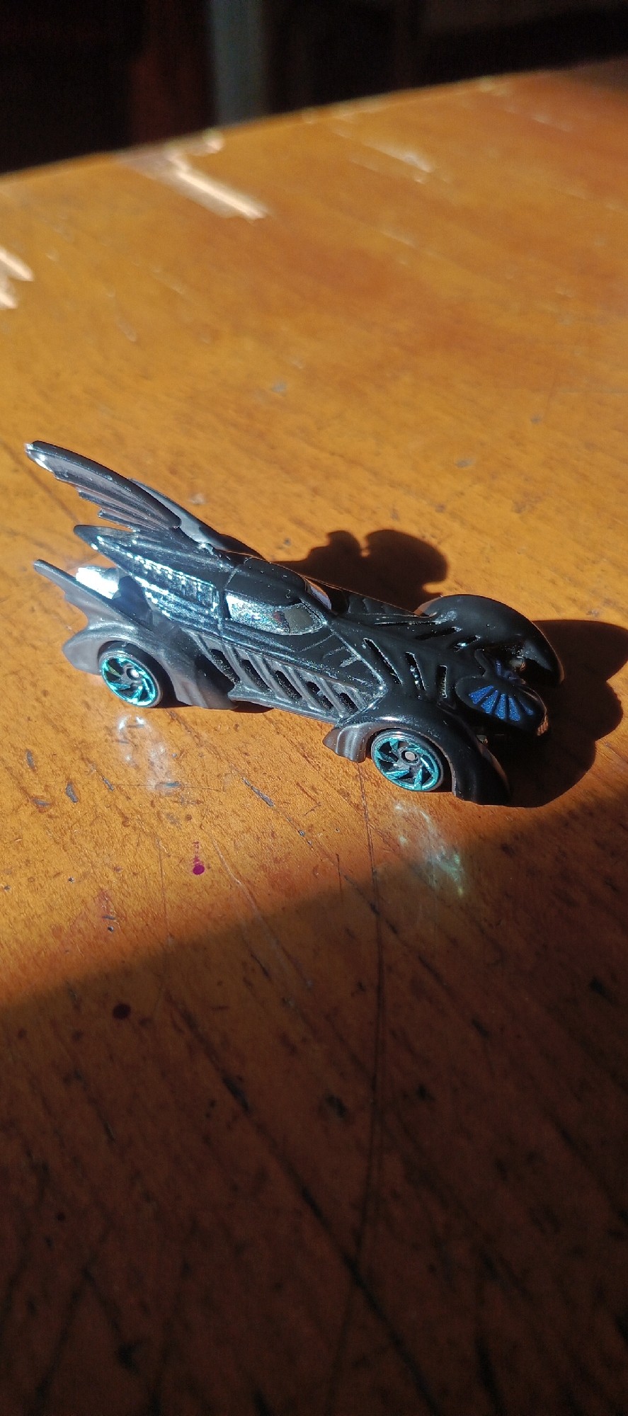 A black Batmobile toy car with blue rims is positioned on a wooden table. The car is in focus and appears to be a Hot Wheels brand toy. The table surface shows wood grain and has a reddish-brown hue. Sunlight casts a strong shadow of the car onto the table. There are some minor scuffs and marks on the table surface. The image is taken from a slightly elevated angle. The scene is not located in Surabaya, Indonesia, but rather in an indoor setting with a wooden table.