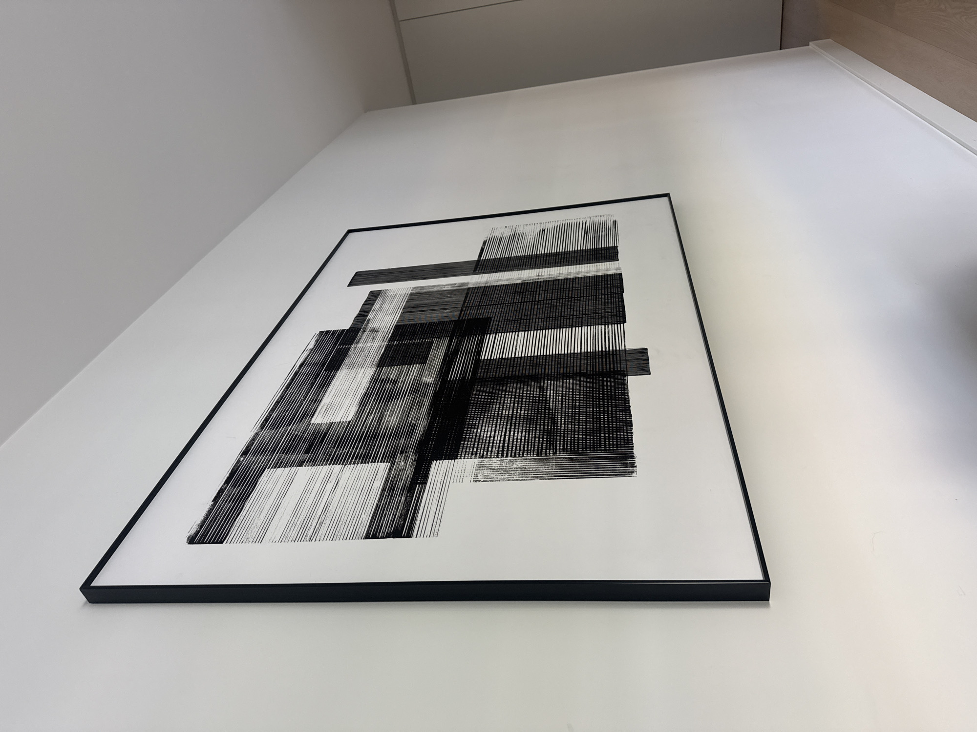 A black and white abstract artwork with a thick black frame hangs on a plain white wall. The artwork is characterized by bold, horizontal and vertical lines, creating a sense of depth and geometric form. The lighting suggests an indoor setting, likely a modern interior. The location is identified as being in the city of Loures, Portugal.