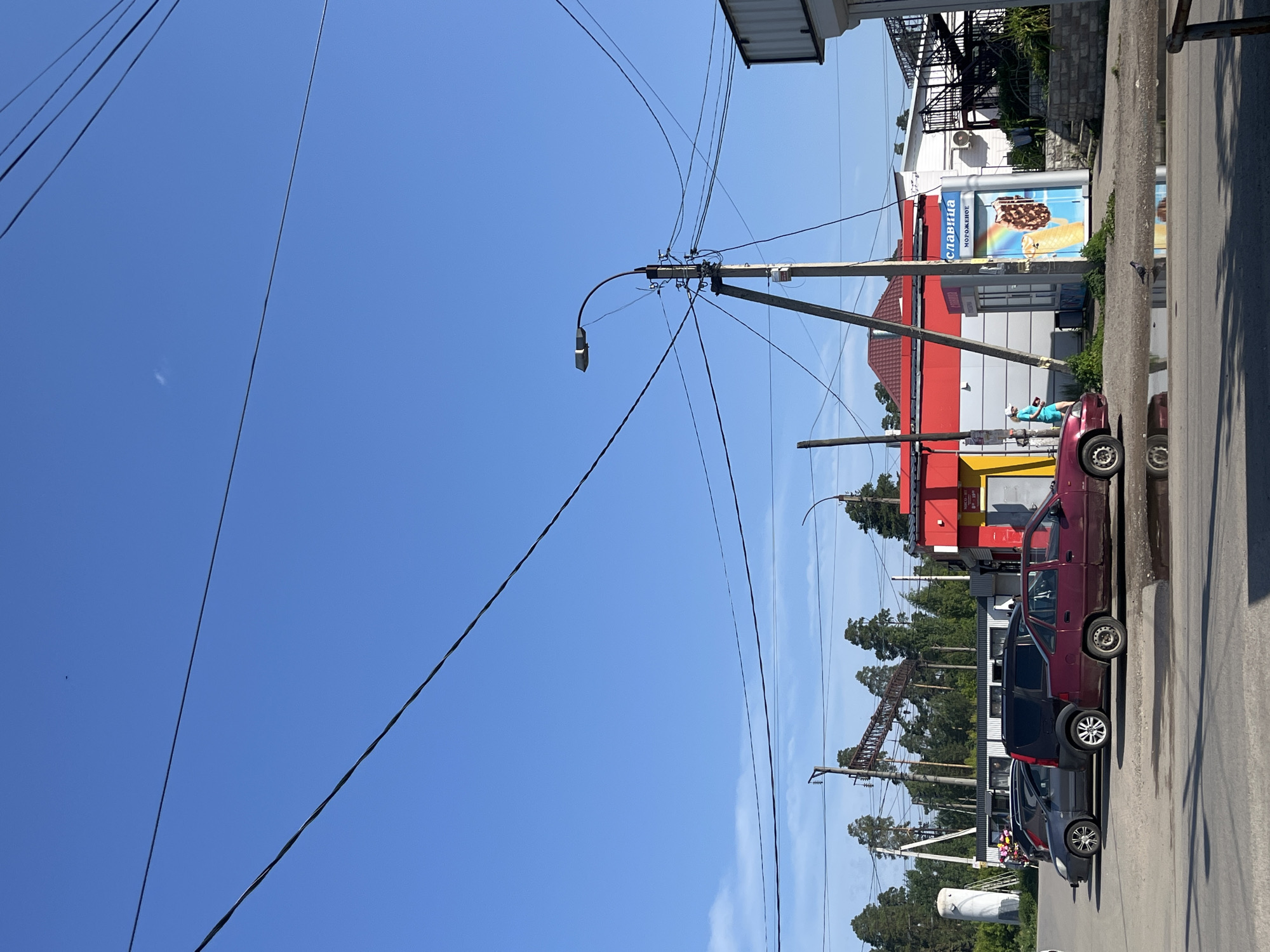 In Vasilyevo, Russia, under a clear blue sky, a street scene unfolds. Power lines crisscross overhead, supported by wooden and concrete poles, one of which features a street lamp. In the foreground, a red car is parked, its tires visible on the asphalt. To its left, a dark SUV and a grey car are also parked.
Behind the parked cars, a row of buildings runs along the street. The prominent structure is a red and white building with signage that includes "СЛАВИЦА" and "МОРОЖЕНОЕ", indicating an ice cream vendor. A woman wearing a blue dress and a white hat walks by the building. Further to the left, a tall white cylindrical structure and a large metal frame with crisscrossing beams suggest industrial or utility infrastructure. Pine trees form a dense backdrop to the scene, with some taller structures visible amidst them. A puddle of water reflects the surrounding environment, with a pigeon standing nearby. The overall impression is one of a functional, everyday urban setting in a locality with abundant greenery.