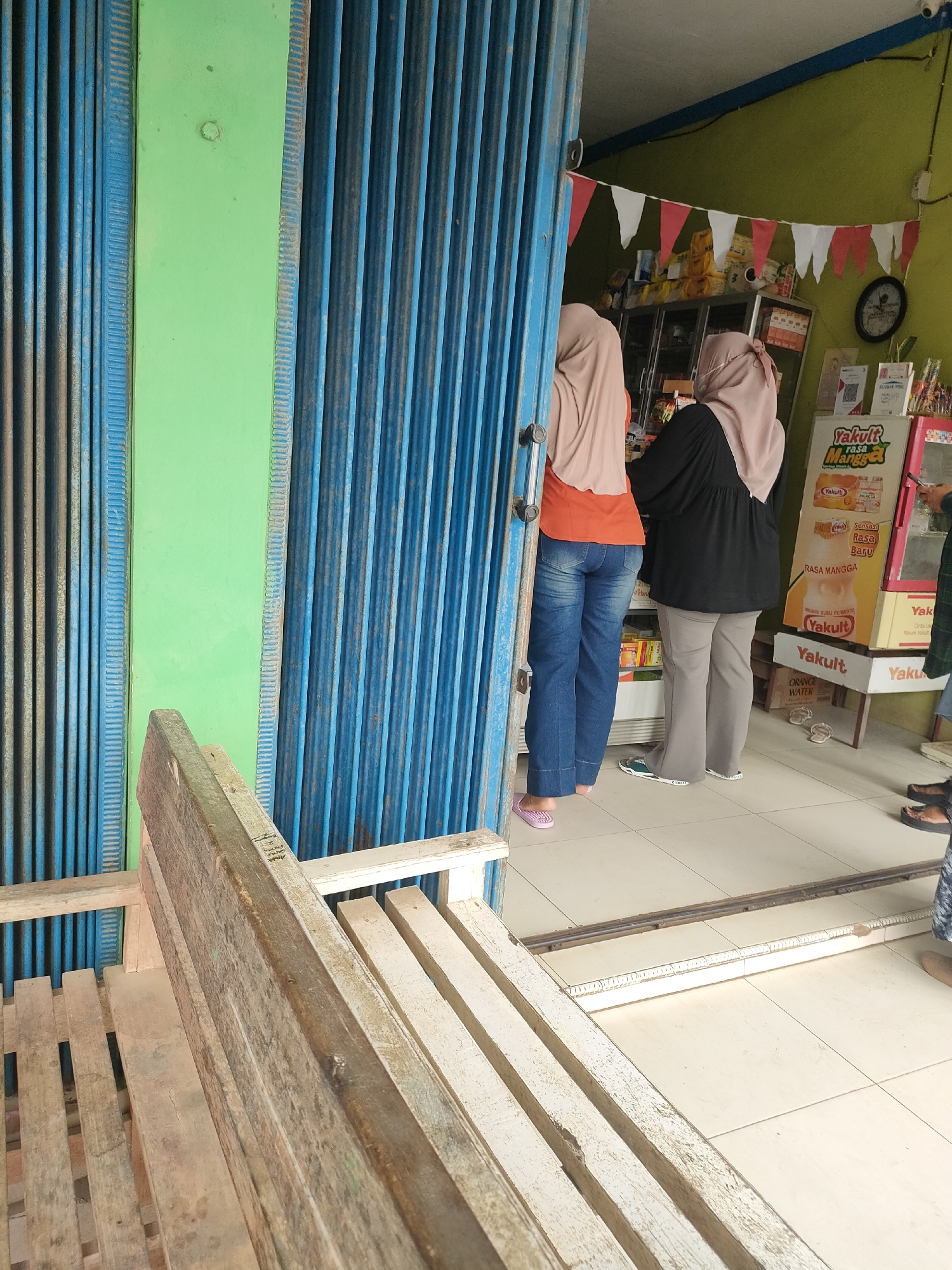 The image depicts the entrance of a small shop or convenience store in Sukoyoso, Indonesia. A weathered wooden bench is positioned in the foreground, partially obscuring the view of the store's interior. Through an open blue metal roll-up door, two individuals are visible inside. One person, wearing a peach-colored hijab and an orange shirt, stands facing away from the camera, wearing blue jeans and pink slippers. To their right, another person in a peach-colored hijab and a black top is also facing away, dressed in gray pants and dark footwear. The interior of the store features shelves stocked with various products, including drinks like Yakult, and a refrigerated display case. Red and white triangular flags are strung across the upper interior wall. A clock is mounted on the green wall behind the customers. The flooring inside the store appears to be tiled. The overall environment suggests a typical neighborhood store.