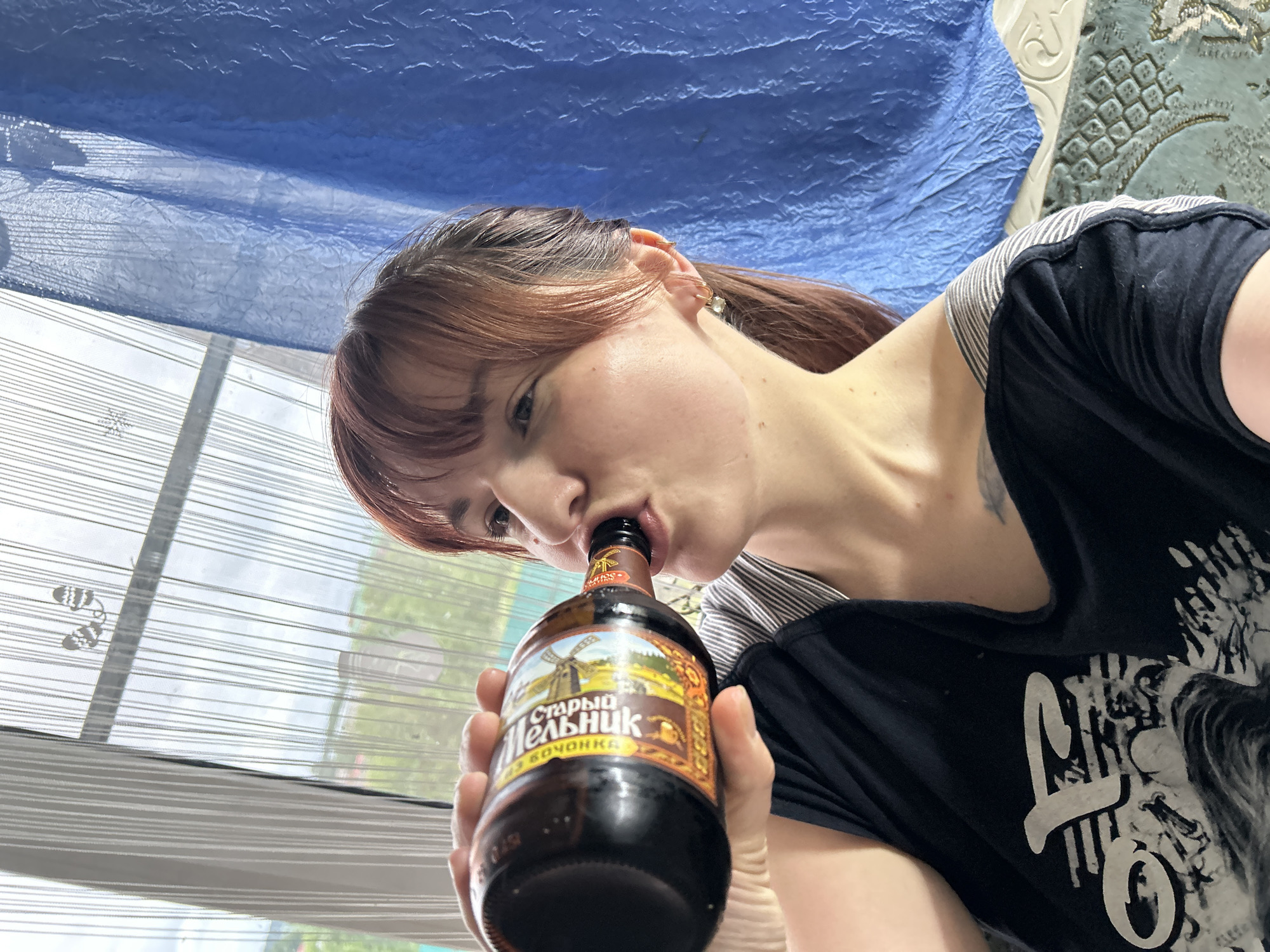 An image captured indoors in the city of Фоки, Russia, depicts a young adult female from the chest up, drinking directly from a dark brown glass bottle. The woman, with reddish-brown hair and bangs, is positioned on the right side of the frame, facing left. Her right hand grips the bottle, which features a colorful label with a windmill design and the Cyrillic text "СТАРЫЙ МЕЛЬНИК". She wears a dark tank top over a striped garment, and a small tattoo is visible on her left upper chest. Her right ear has a small hoop earring with a pearl and a cartilage piercing. The background features a large window covered by a sheer white vertical string curtain, through which green trees and distant buildings are visible under an overcast sky. A small snowflake decal and two mitten-shaped decorations are affixed to the sheer curtain. A blue, textured opaque curtain is partially drawn on the right. In the lower right foreground, a section of an ornate light-colored furniture piece and a patterned rug with blue and green tones are visible.