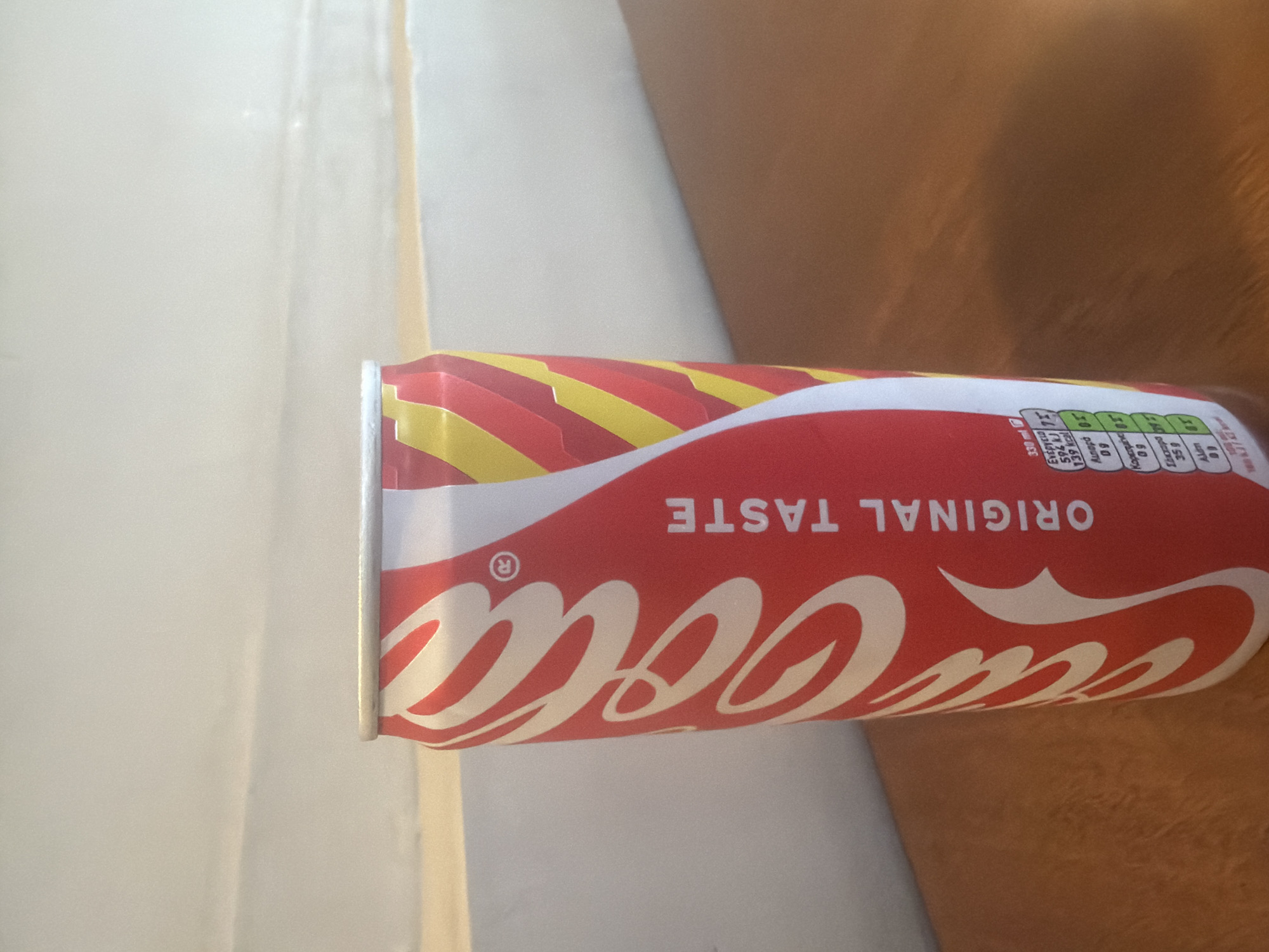 A close-up, vertically oriented shot shows a red Coca-Cola can lying on its side, partially obscuring a blurred background that suggests an indoor setting, possibly a bathroom, with a white tiled surface on the left and a warm, skin-toned surface on the right. The can is the primary subject, with its iconic red and white branding prominently displayed. The "Coca-Cola" logo is clearly visible in its classic script font, curving along the body of the can. Above the logo, the words "ORIGINAL TASTE" are printed in white capital letters. The top of the can features a yellow and red striped design, with a silver pull-tab visible.

On the right side of the can, a section of the nutrition facts label is visible. The text is in a small font and appears to be in Greek, with terms like "Ενέργεια" (Energy), "Θερμίδες" (Calories), "Λιπαρά" (Fat), "Κορεσμένα" (Saturated), and "Αλάτι" (Salt) readable. The numbers indicate values such as 594 kJ, 139 kcal, and 0.9g for fat and saturated fat.

The lighting in the image is soft, casting gentle shadows on the right side of the can and highlighting the texture of the skin-toned surface. The overall impression is a still life focusing on the beverage product, with the background elements intentionally out of focus. There are no people clearly visible, and the scene is static. The location context of Kiti, Cyprus, is noted, but no specific visual cues within the image directly confirm this location.