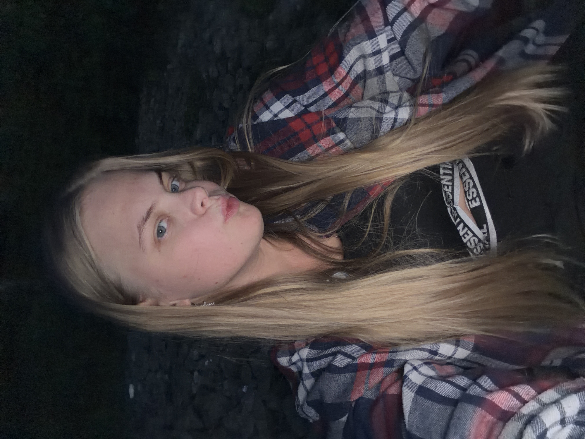 A young woman with blonde hair is facing the camera with puckered lips. She is wearing a black top with white lettering visible on the strap. A plaid shirt with red, white, and blue checks is draped around her. The background is dark, suggesting an outdoor setting, possibly a rocky surface on the left. Without additional information, determining the location as Izhevsk, Russia, is not possible.