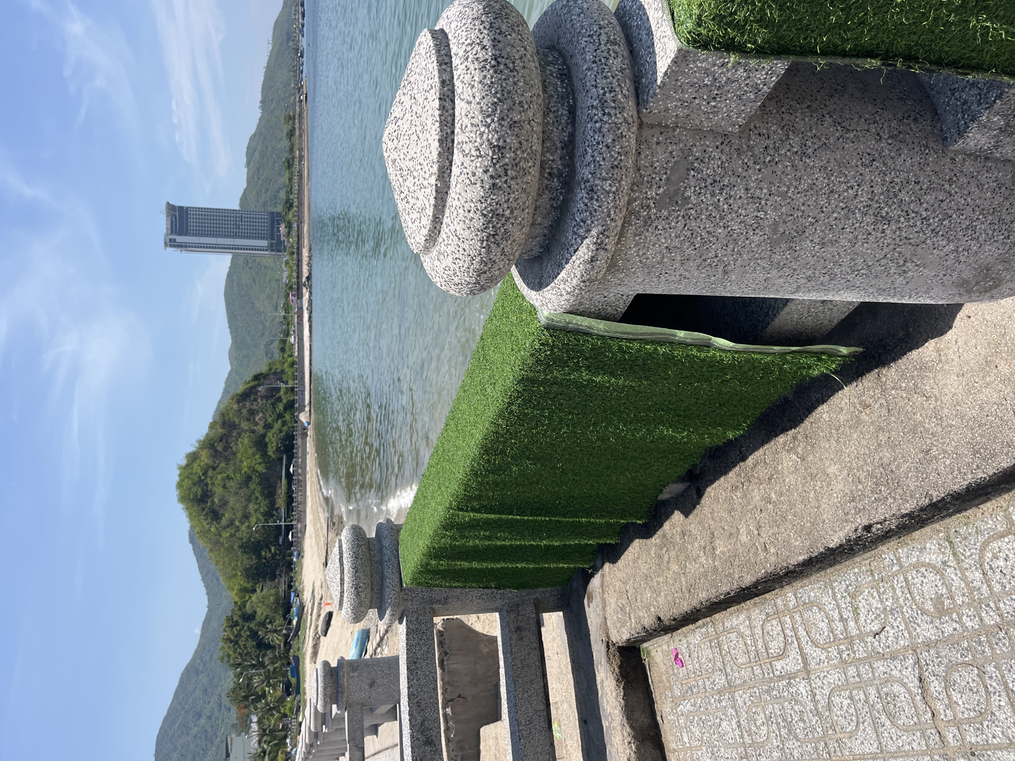This image captures a sunlit coastal scene in Bac Nha Trang Ward, Vietnam. In the foreground, a grey, speckled concrete balustrade extends diagonally across the lower portion of the frame, featuring urn-shaped finials atop its posts. Artificial green turf is draped over the balustrade's horizontal rail, covering its seaward side. To the left of the balustrade, a patterned concrete walkway exhibits a geometric motif, with a single pink flip-flop resting on its surface. An elevated concrete curb runs parallel to the walkway. Strong shadows from the balustrade and turf are cast onto the concrete, indicating direct sunlight.
The midground reveals the calm, blue-green water of a bay, leading to a sandy beach. Several small boats, some blue, are visible on the sand or at the water's edge. A line of green foliage, including palm trees, borders the beach, behind which a tree-covered hill rises.
The background features a range of green, forested mountains under a clear, bright blue sky with sparse white clouds. A tall, slender grey skyscraper, exhibiting numerous windows and appearing under construction at its summit, stands prominently on the right side, situated between the mountains and the water. No individuals are visible in the scene.