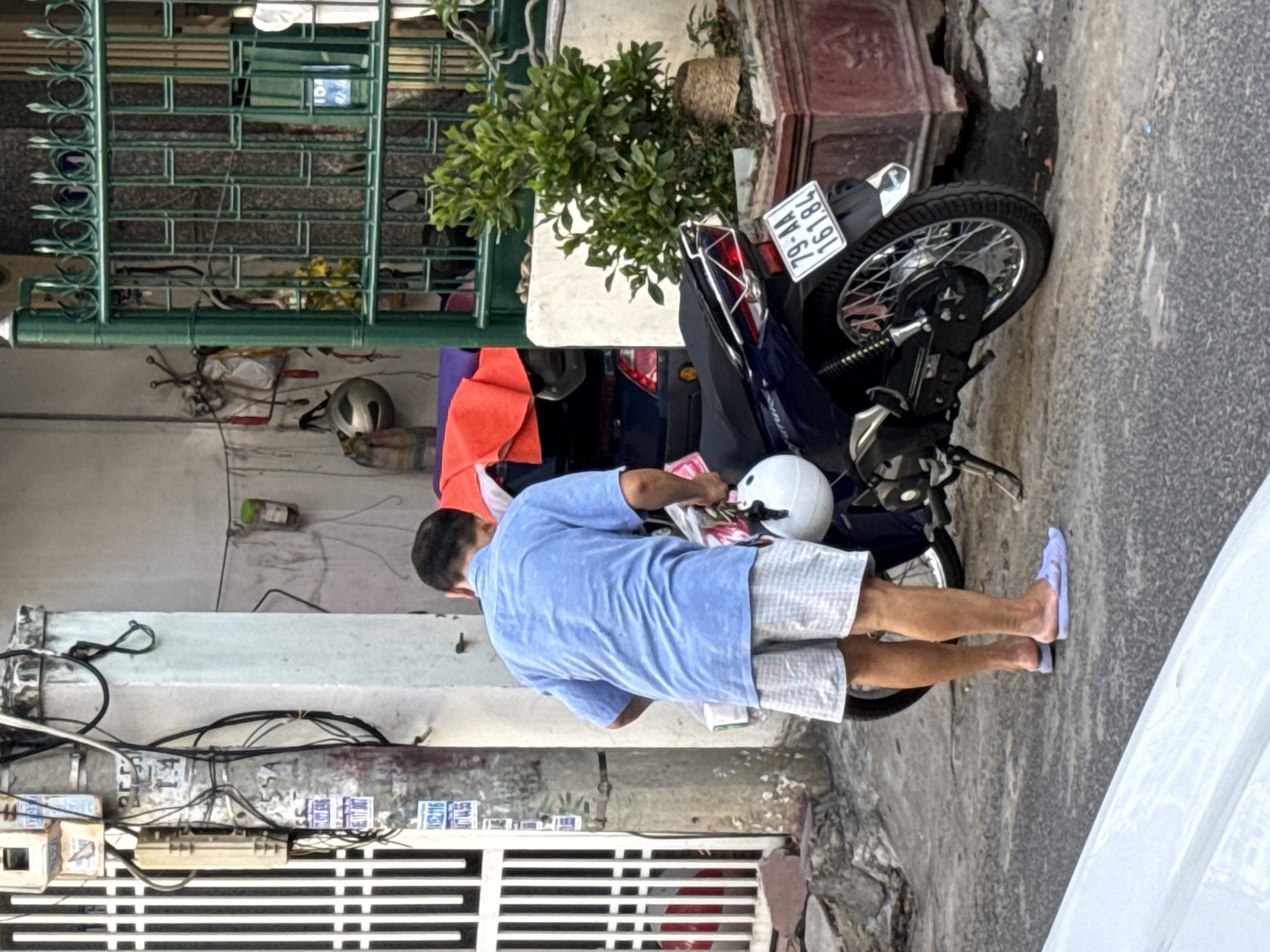 A man in a light blue short-sleeved shirt and checkered shorts stands next to a dark blue motorcycle parked on a paved street in Nha Trang, Vietnam. He is holding a white helmet in his right hand and appears to be preparing to mount the motorcycle. The motorcycle has a license plate that reads "79-AA 161.84". In the background, there is a green metal gate, a potted plant, electrical poles with wires, and a white building. The scene is outdoors, likely in a residential or commercial area, with evidence of daily urban life.