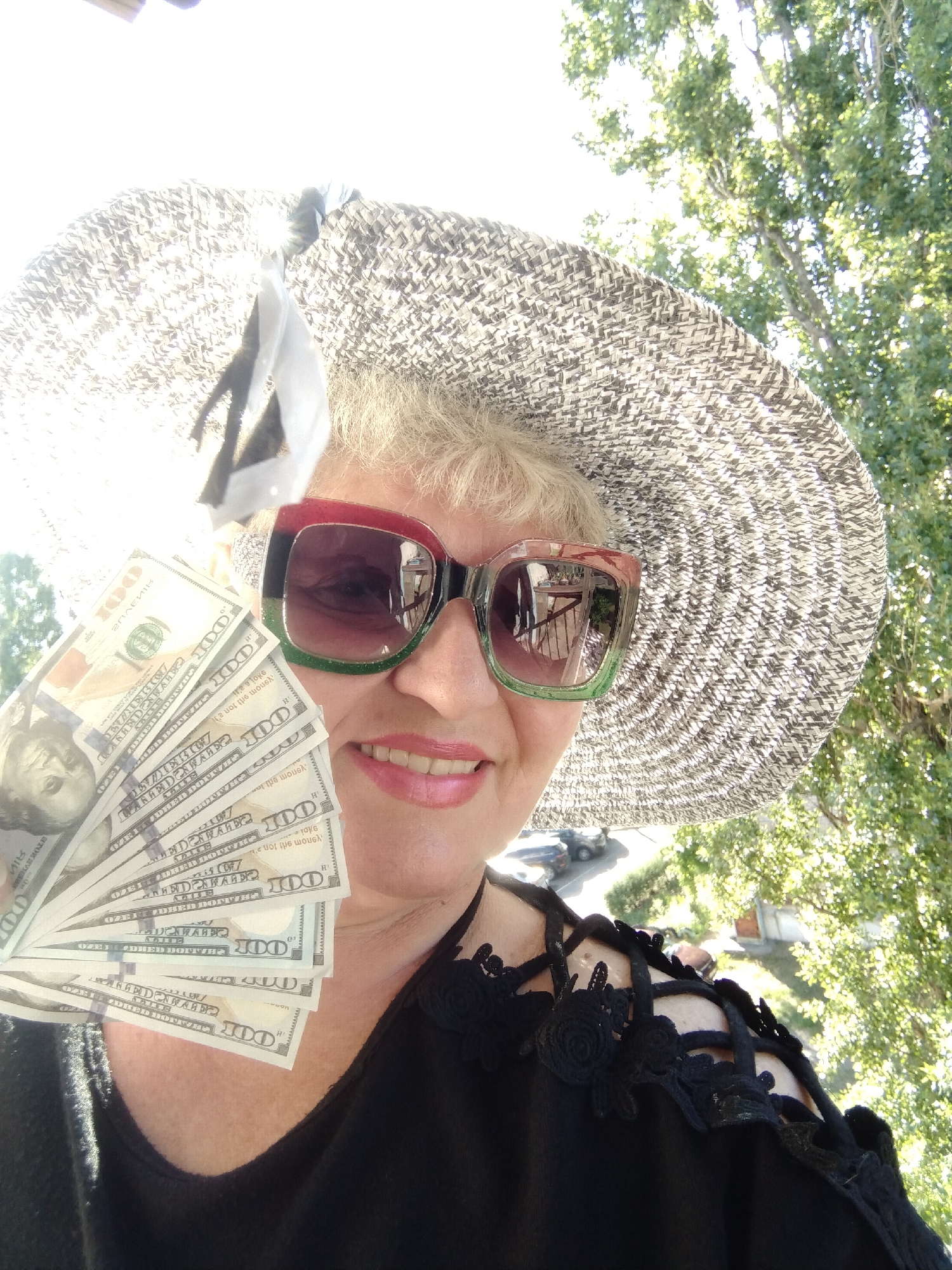 A blonde woman wearing a large straw hat and colorful sunglasses holds a fan of US hundred-dollar bills in her right hand, smiling at the camera. She is wearing a black top with decorative straps. The background shows green foliage and sunlight, suggesting an outdoor setting. The provided crops suggest the image was taken from an elevated position, possibly a balcony, overlooking a street with cars. The location of Horishni Plavni, Ukraine is not discernible from the image itself.