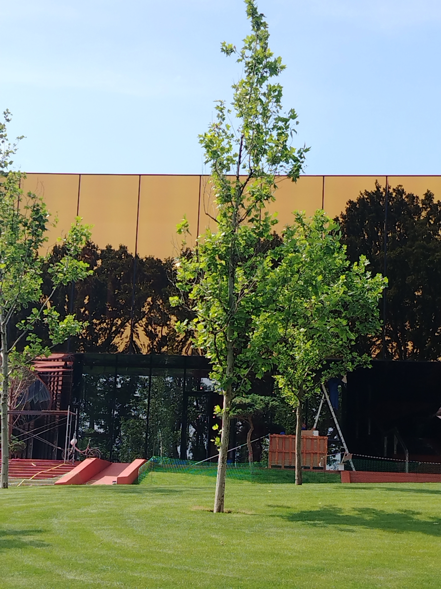 The image depicts a modern building with a large, reflective, golden-orange facade, likely a commercial or public structure, situated in an urban park setting. The facade reflects an image of dense trees. In the foreground, a meticulously manicured green lawn is visible, with several young trees strategically planted. To the left, a set of red ramps or slides are installed on the lawn, with scaffolding and construction materials nearby, suggesting ongoing development or renovation. A pink bicycle with a white basket is parked on top of one of the ramps. In the mid-ground, the reflective building is prominent, with its clean lines and modern design. The sky above is clear and blue. The overall scene conveys a sense of contemporary architecture integrated with green spaces, possibly a park or a recreational area within the city of Krasnodar, Russia.