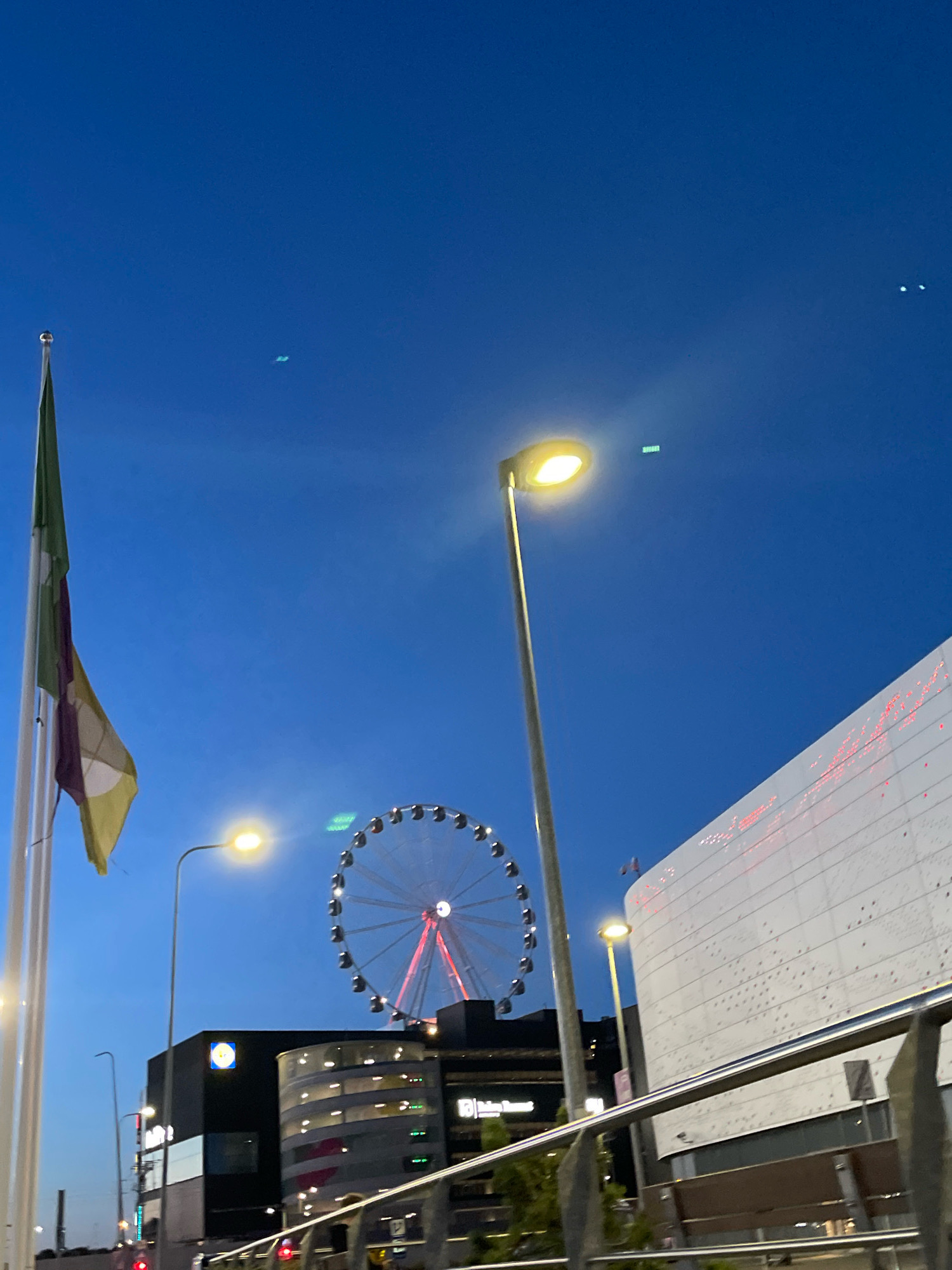 The image depicts an evening scene in Tallinn, Estonia, featuring a large Ferris wheel as the central element. To the left, a flagstaff with a flag is visible. A lamppost with a lit bulb stands in the foreground. Behind the Ferris wheel, a modern building with a white facade and red lighting accents can be seen. To the left of the Ferris wheel, a dark building with illuminated signage is present. The sky is a deep blue, characteristic of twilight or early night. The overall impression is that of an urban entertainment or leisure area.
