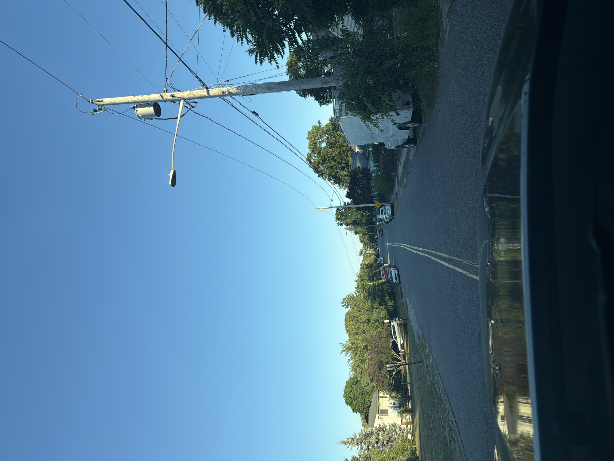 The image shows a residential street in Riverhead, New York, USA, on a clear day with a bright blue sky. The perspective is from the driver's seat of a vehicle, looking forward down a paved road with a double yellow line in the center. On the left side of the road, there are houses with lawns and trees, and several parked cars. On the right side of the road, there is a utility pole with wires and a street lamp, as well as lush green bushes and trees. Further down the street, more houses and parked vehicles are visible. A white utility truck is parked on the right side of the road. The overall scene depicts a quiet suburban street.