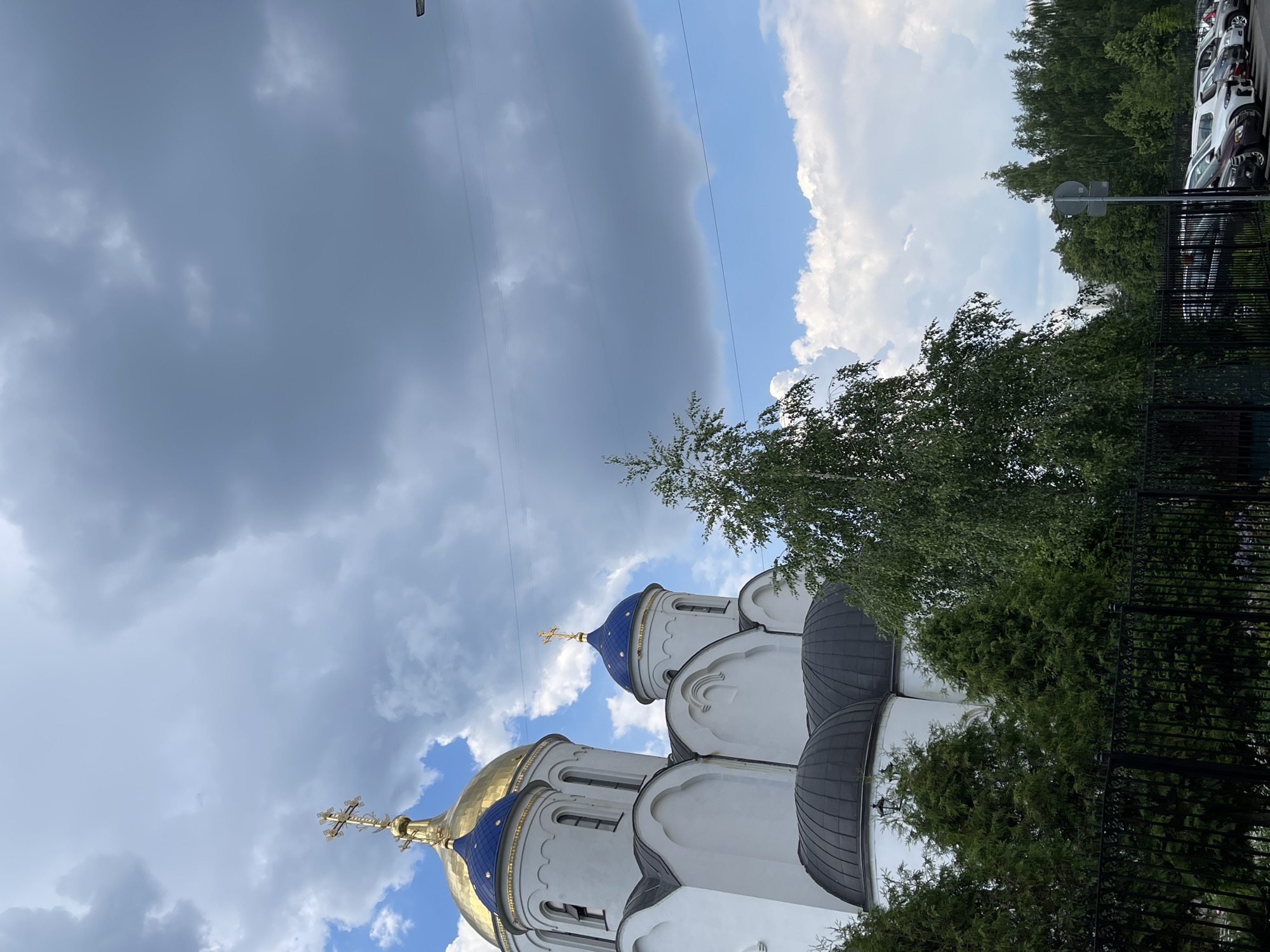 The image shows the Church of the Resurrection of Christ in Tverskaya Zastava Square, Moscow, Russia. The church features white walls and multiple domes, some of which are gilded and blue. A large green tree stands in the foreground, partially obscuring the church. In the background, a row of parked cars is visible, along with a dense forest. The sky is partly cloudy, with dramatic dark clouds dominating the upper portion of the frame and bright blue sky with white clouds visible in the lower portion. There are no people visible in the image.
