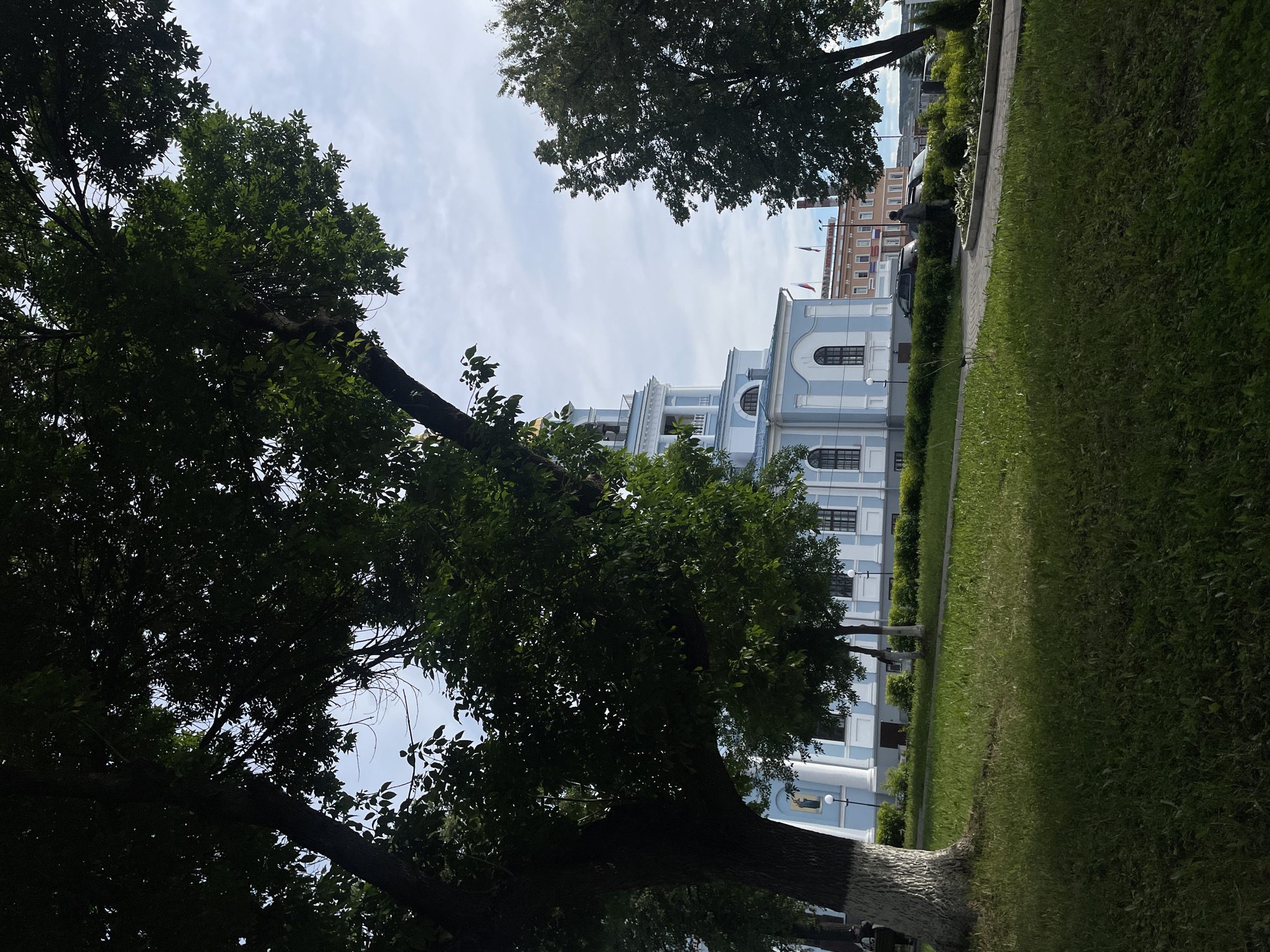 A serene outdoor scene on a clear, bright day, likely in Votkinsk, Russia. The foreground and sides are dominated by the dark green foliage of large trees, creating a natural frame. Through the trees, a grand light blue and white building stands prominently in the mid-ground. It features classical architecture with arched windows, white columns, and a multi-tiered tower or cupola.
To the right of the building, a wide expanse of neatly manicured green grass stretches alongside a paved pathway. Low hedges and streetlights line the path in front of the building. Further in the background, a multi-story building with brown and orange facades is visible. On this distant building, Cyrillic text reading "МЕБЕЛЬ" (Furniture) and "ТЕЛЕФОН" (Telephone) can be discerned. Russian flags are seen flying from poles on both the main blue building and the distant brown building.
The sky is bright with scattered white clouds, suggesting daytime, possibly late morning or early afternoon. No explicit activity is taking place, presenting a peaceful and calm atmosphere. A small, dark figure, potentially a statue or person, is faintly visible on the distant pathway, and several parked cars are behind the buildings in the background.