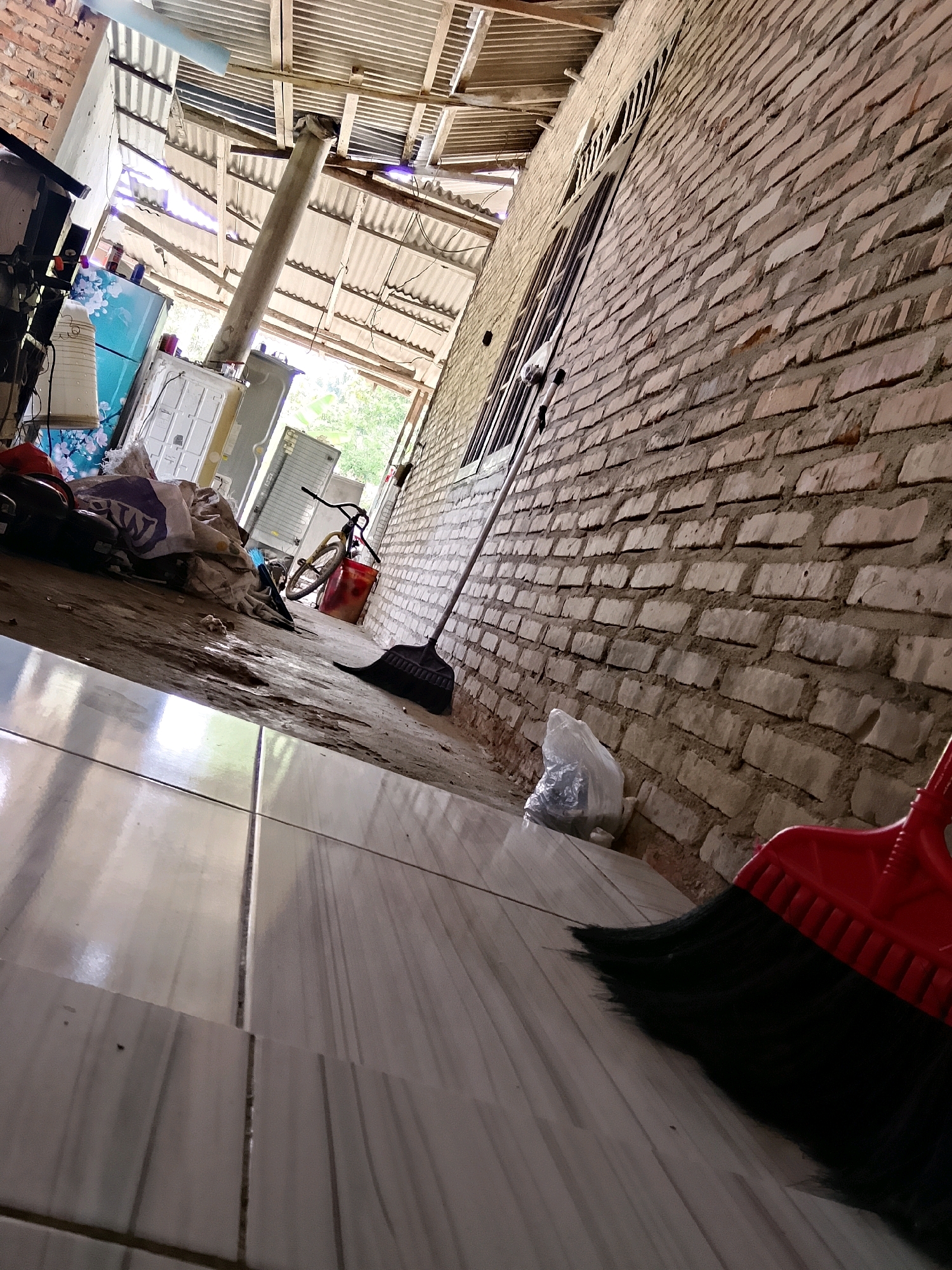 The image depicts an outdoor scene with a brick wall on the right and a corrugated metal roof overhead. The ground is a mix of dirt and tiled flooring. In the foreground, a red broom with black bristles is visible. To the left, there is a pile of items including a blue and white patterned refrigerator, a white appliance, a bicycle with a red bucket, and several bags of what appear to be trash or debris. A black broom is also present, leaning against a window. The environment suggests a somewhat unkempt outdoor area, possibly a backyard or a service alley in Karang Gading, Indonesia, characterized by its rustic construction and the presence of discarded items.