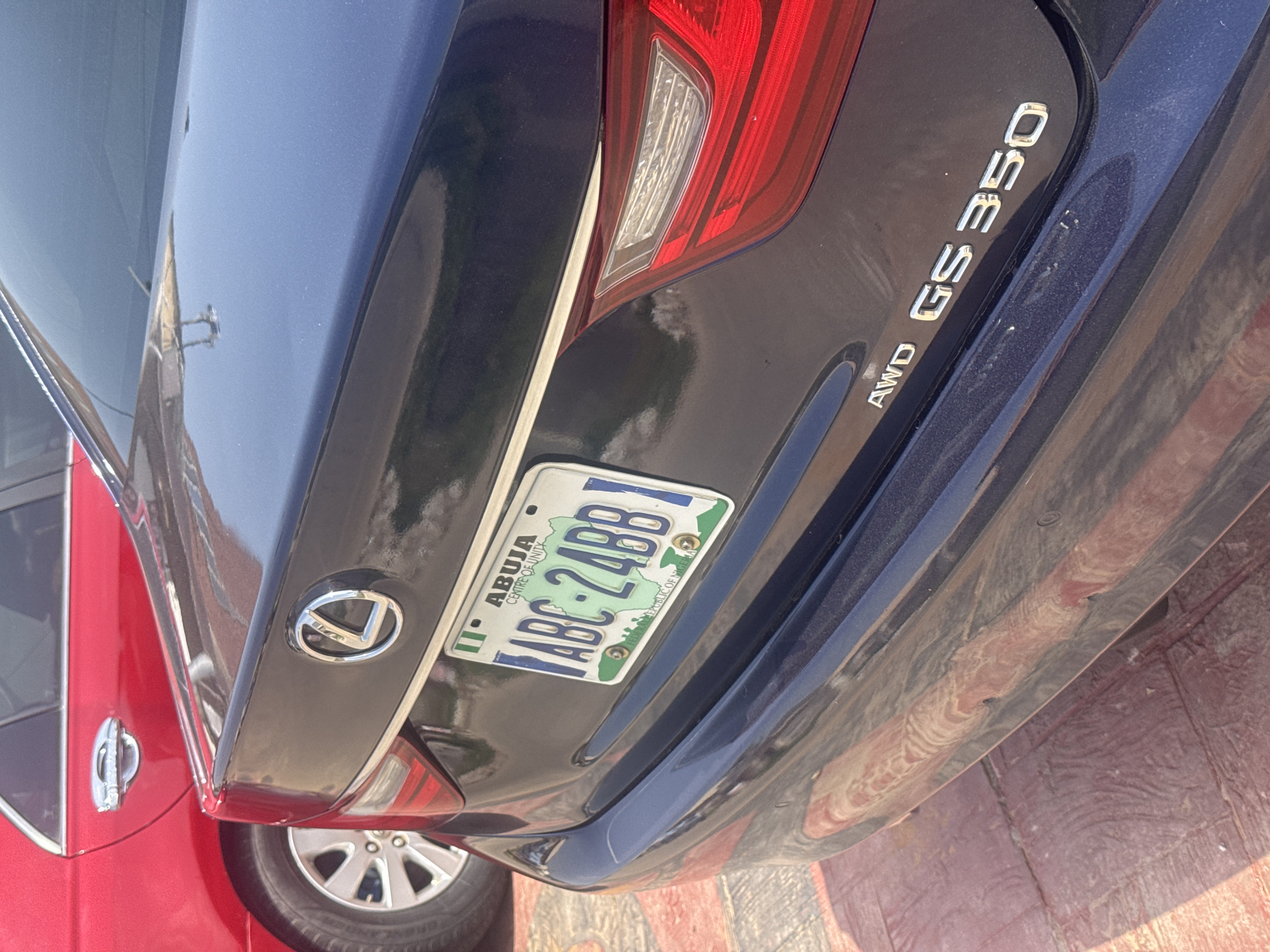 This media file shows the rear of a dark-colored Lexus GS 350 vehicle parked outdoors. The car is positioned at an angle, with its taillight and license plate prominently displayed. The license plate, from Abuja, Nigeria, reads "ABC 24BB" and also displays "CENTRE OF UNITY" and "FEDERAL REPUBLIC OF NIGERIA". The car's emblem, a stylized "L", is visible on the trunk.
To the left of the Lexus, a portion of a red car is visible, featuring a chrome door handle. The setting appears to be a paved area with some textured, possibly reddish, ground covering. The lighting suggests it is daytime, with the sun casting reflections on the car's glossy surface. No people are visible in the image. The overall scene is a static display of two vehicles.