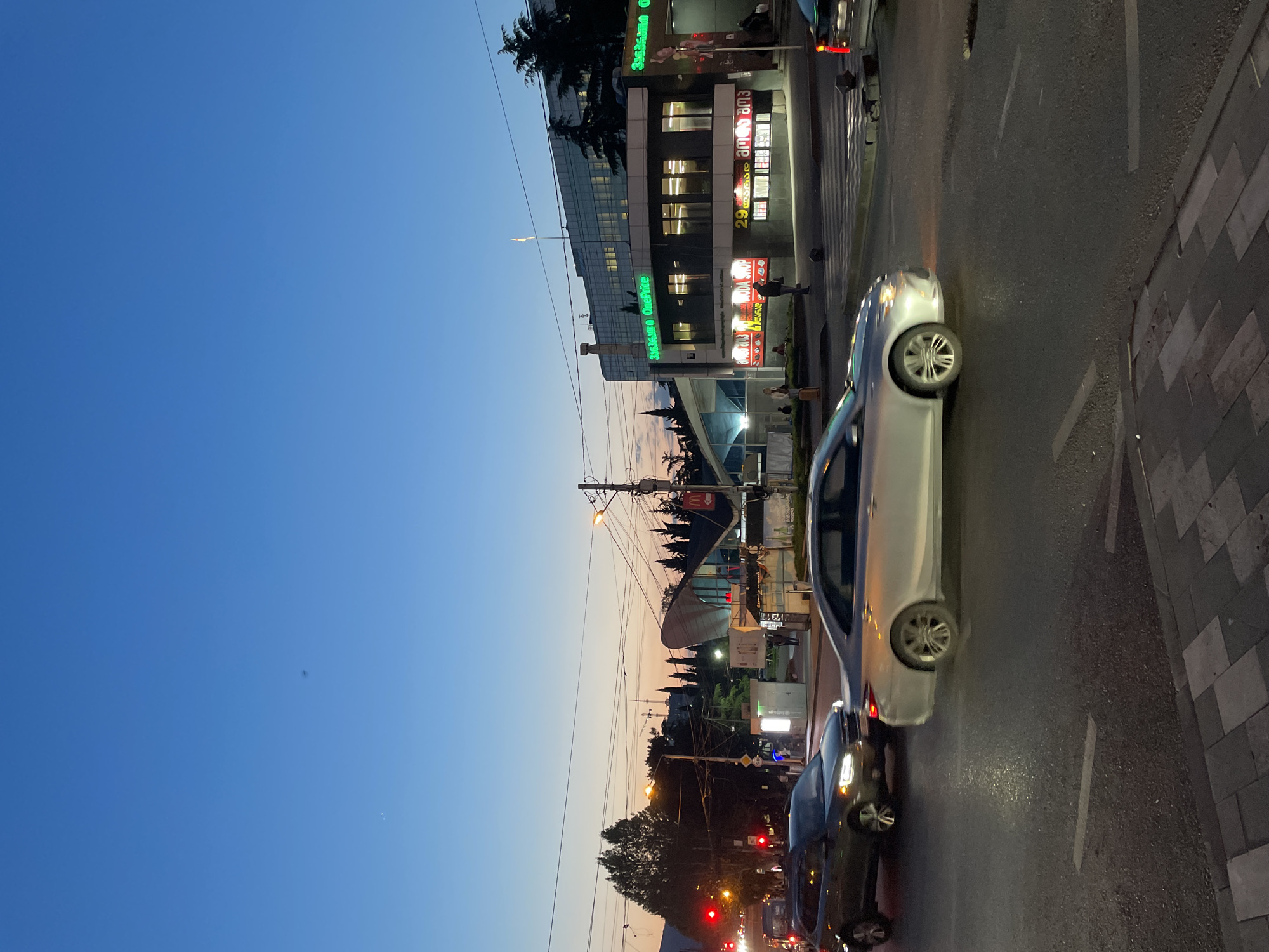 This image captures an urban street scene during dusk or early evening in Tbilisi, Georgia. The sky, visible at the top, transitions from a deep blue to a lighter, warmer orange-yellow gradient near the horizon, indicative of twilight.
In the foreground and mid-ground, an asphalt road features white lane markings. Several vehicles are present, including a prominent light-colored sedan and a darker car following behind it. A paved sidewalk with square tiles runs along the right side of the road.
On the left side of the street, multiple commercial buildings line the view. A modern building with reflective glass facades and illuminated windows stands prominently. Below it, several storefronts glow with bright lighting. Green neon signs display "Sushiroom OnePrice" and "Sushiroom." Other signs feature text in Georgian script, with "ამბი" (Ambi) and the number "29" clearly discernible. A red "M" logo, typical of McDonald's, is also visible on one of the signs.
The scene includes utility poles with numerous overhead wires, and streetlights are beginning to illuminate the area. In the distance, a large dish-like structure and what appears to be a construction crane can be seen against the evening sky. Trees are scattered throughout the urban landscape. A few indistinct human figures are visible walking on the sidewalk, adding to the sense of routine city activity. The image conveys the quiet bustle of a city transitioning from day to night.
