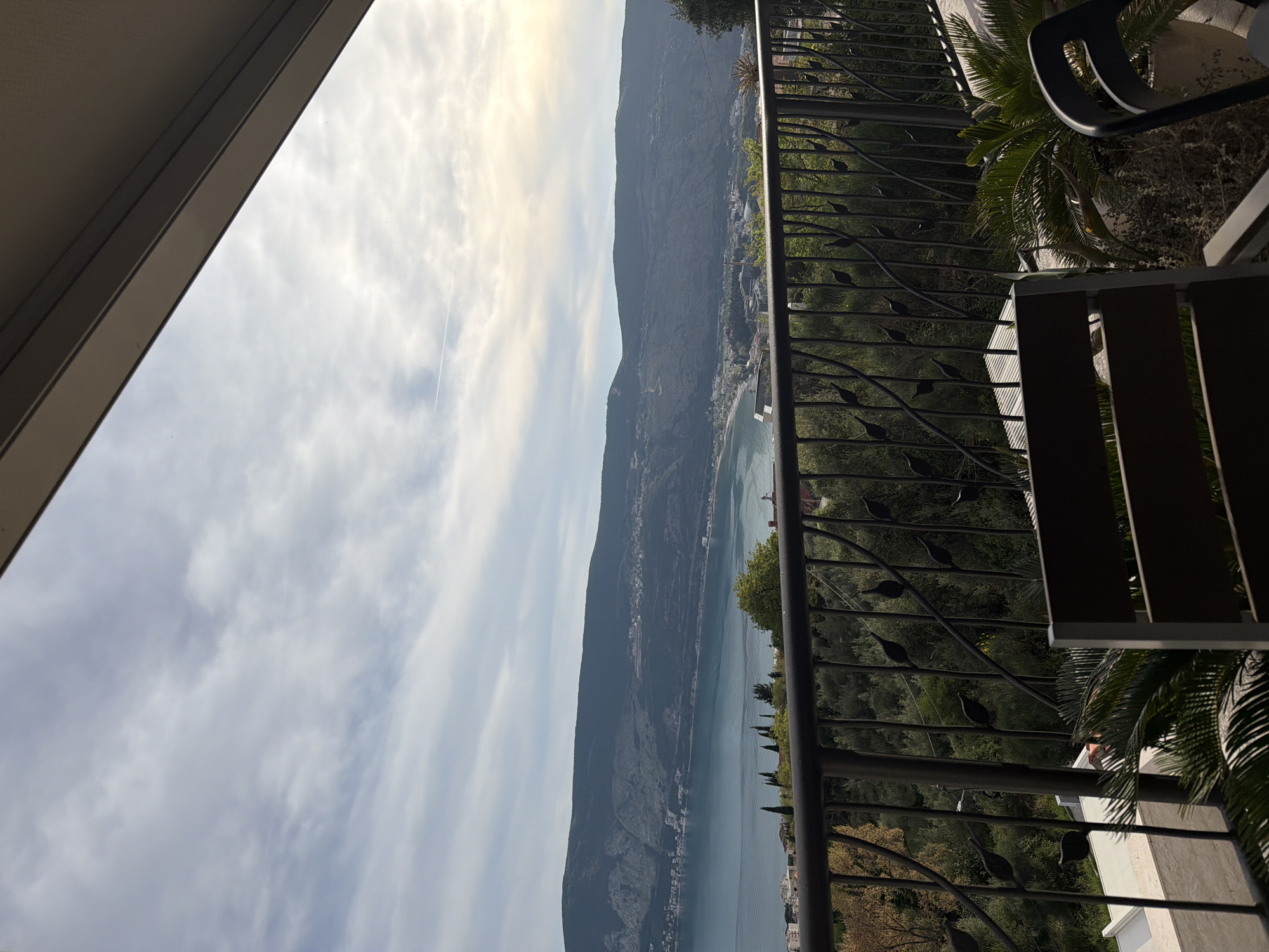 View from a balcony in Trebesin, Montenegro. The balcony has a decorative metal railing and furniture. Beyond the railing, there is a landscape featuring a body of water with a mountain range extending along its shore. A city is visible nestled between the water and the mountains. The sky is cloudy with visible sunlight. The image appears to be taken during daylight hours.