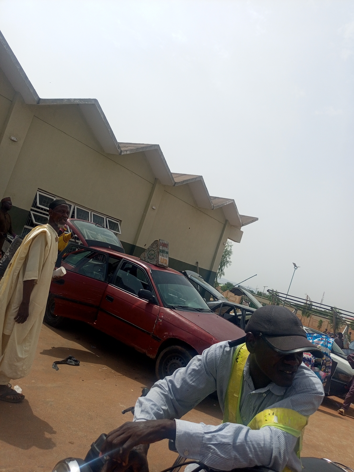 This image captures a vibrant outdoor scene, likely a transport hub or market area, under a bright sky in Gombe, Nigeria.
In the immediate foreground, a man on a motorcycle is visible from the chest up. He wears a light blue and white striped shirt, a dark baseball cap, and bright yellow reflective bands on his arms. He is looking slightly to his right with a faint smile, his hands gripping the motorcycle handlebars.
To the left, a man in a long, cream-colored robe (agbada-like) and a dark cap stands near a maroon hatchback car, holding a white cup. He has a beard and appears to be observing the scene. The maroon car, showing signs of wear, has its trunk open. A prominent sign on its roof displays the words "GOMBE," "DUKKA," and "RAYA," along with other less distinct text, suggesting it is a taxi or commercial transport vehicle. Another person is partially visible behind the open trunk of the car.
The background is dominated by a light-colored building featuring a distinctive jagged or zigzag roofline and multiple rectangular windows. Other vehicles are parked further behind the maroon car, and a solar-powered street light stands in the distance. The ground beneath is unpaved, appearing as dirt or sand. The bright, clear sky and strong shadows indicate a sunny day, likely midday or early afternoon.