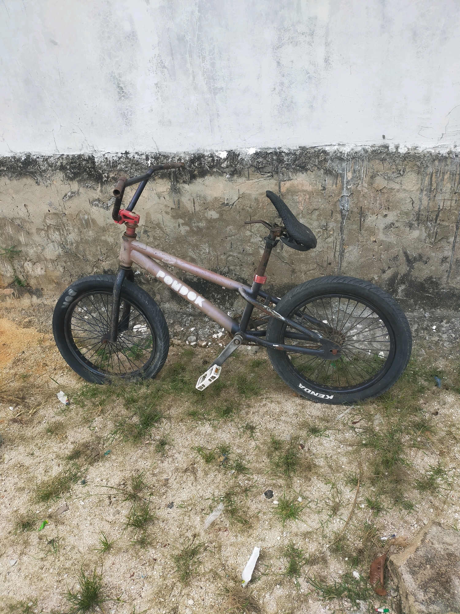 A BMX bicycle is parked outdoors against a weathered concrete wall. The bicycle has a metallic brown frame with white lettering that appears to read "PONSOK" on the top tube. It features black tires, one of which has "KENDA" written on the sidewall in white. The bicycle has a black saddle and pedals. The ground is a mixture of sand and sparse grass, with some scattered debris. The wall behind the bicycle shows signs of age and wear, with patches of peeling paint and dirt. The overall environment suggests an outdoor urban setting, possibly in Pekanbaru, Indonesia, given the context of the request. There are no people visible in the image.