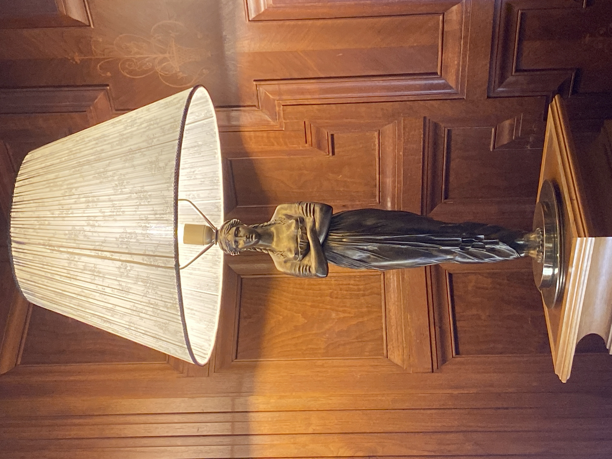 The image depicts a lamp with a statue of a female figure at its base, set against a backdrop of ornate wooden paneling. The statue, made of a dark material, likely bronze, is of a woman with her arms crossed, wearing a draped garment. The lamp shade is light-colored and pleated, with a subtle floral pattern. The wooden paneling exhibits a warm, rich tone and features decorative carvings and recessed sections. The setting appears to be an interior, possibly a room in a historic building, given the elaborate woodwork. The lighting suggests the lamp is illuminated, casting a soft glow. The location in Saint Petersburg, Russia, is not directly discernible from the image itself, but if this were part of a larger context, it would be a descriptive detail of the scene.
