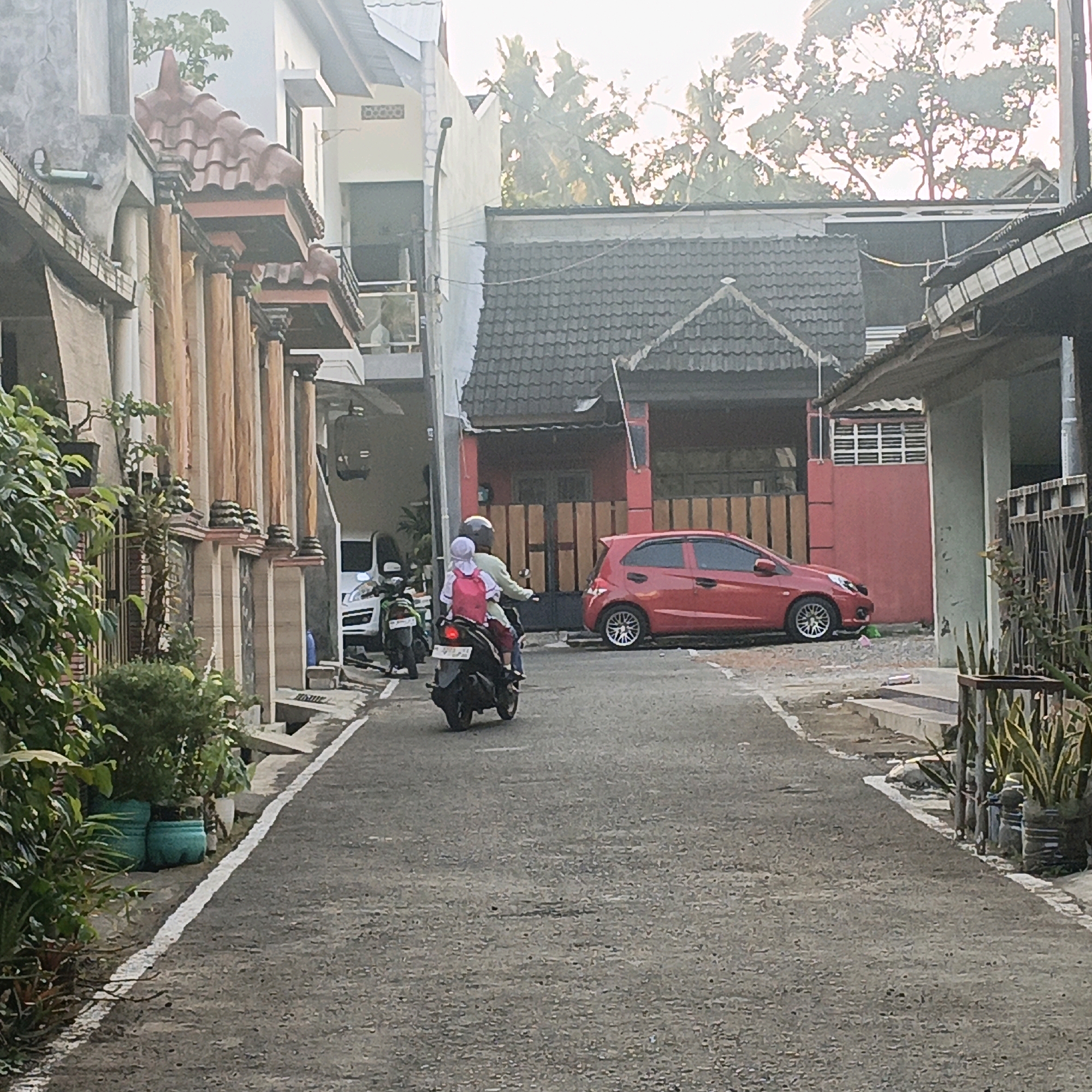 In a residential street in Salatiga, Indonesia, a person drives a motorcycle with a child passenger. The driver, wearing a light green long-sleeved shirt and a helmet, is in the foreground. The child, wearing a white hijab and a pink backpack, is seated behind the driver. The motorcycle is black and moving forward. To the right of the motorcycle, a red hatchback car is parked. The street is paved with asphalt and has a white line marking the edge. On the left side of the street, there is a building with decorative columns and potted plants. On the right side, there is a fence and more potted plants. The background shows houses with tiled roofs and trees under a bright sky. The scene appears to be captured during the daytime, likely in the late afternoon given the lighting conditions.