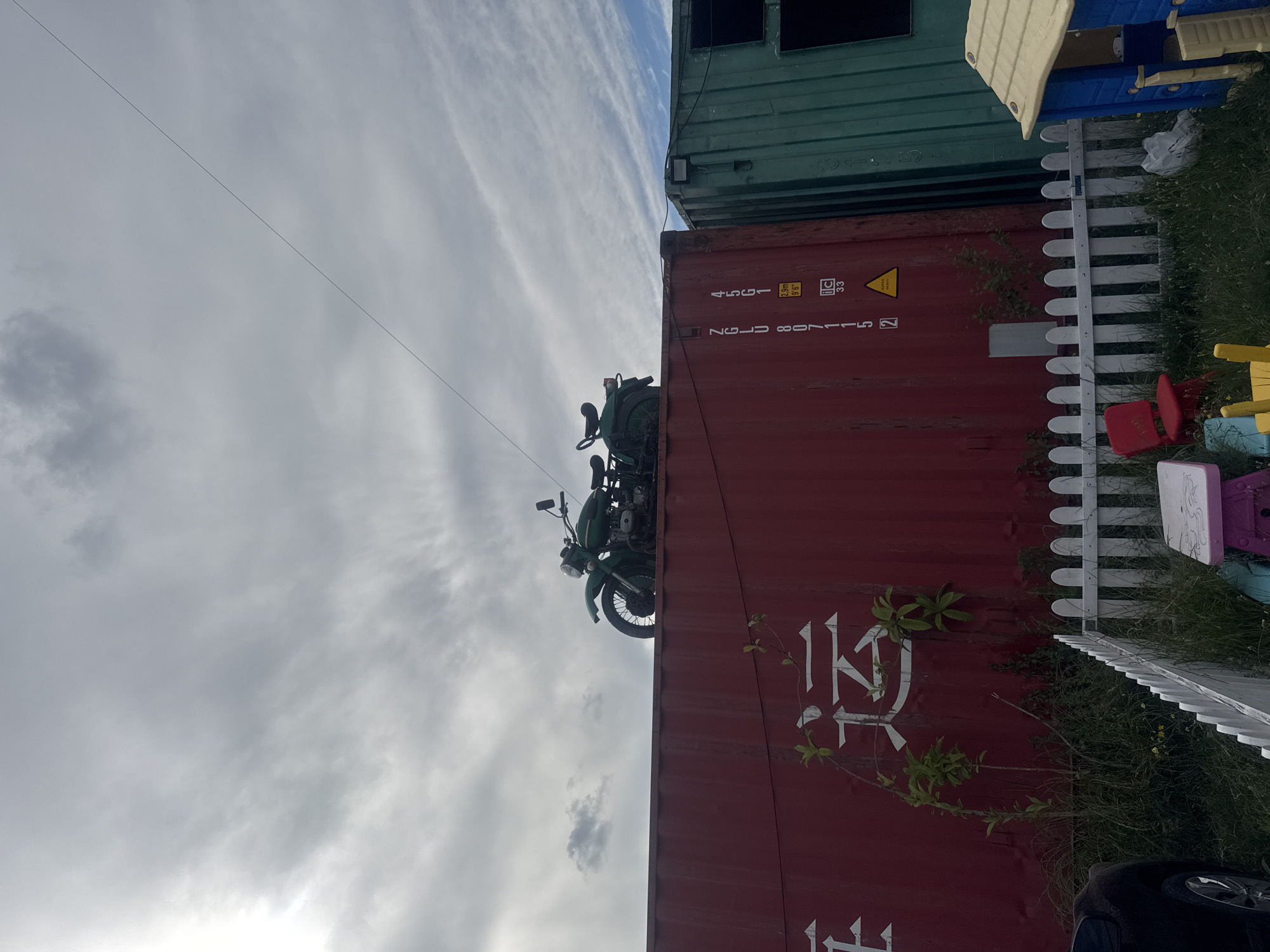 The image depicts a scene outdoors, possibly in a junkyard or storage facility, with shipping containers dominating the frame. A vintage green motorcycle is perched precariously on top of a large red shipping container. The container has white Chinese characters painted on its side, which appear to translate to "transport" or "shipping." Adjacent to the red container is a green shipping container. In the foreground, a white picket fence separates the containers from a grassy area with several children's toys, including a red plastic chair, a small table, and a blue and yellow playhouse. The sky above is overcast with dramatic clouds. While the image does not explicitly state its location, the presence of shipping containers and an outdoor storage environment is consistent with industrial or fringe areas within a city. The general aesthetic does not strongly suggest a specific location within Tbilisi, Georgia, without further contextual information. No people are visible in the image.