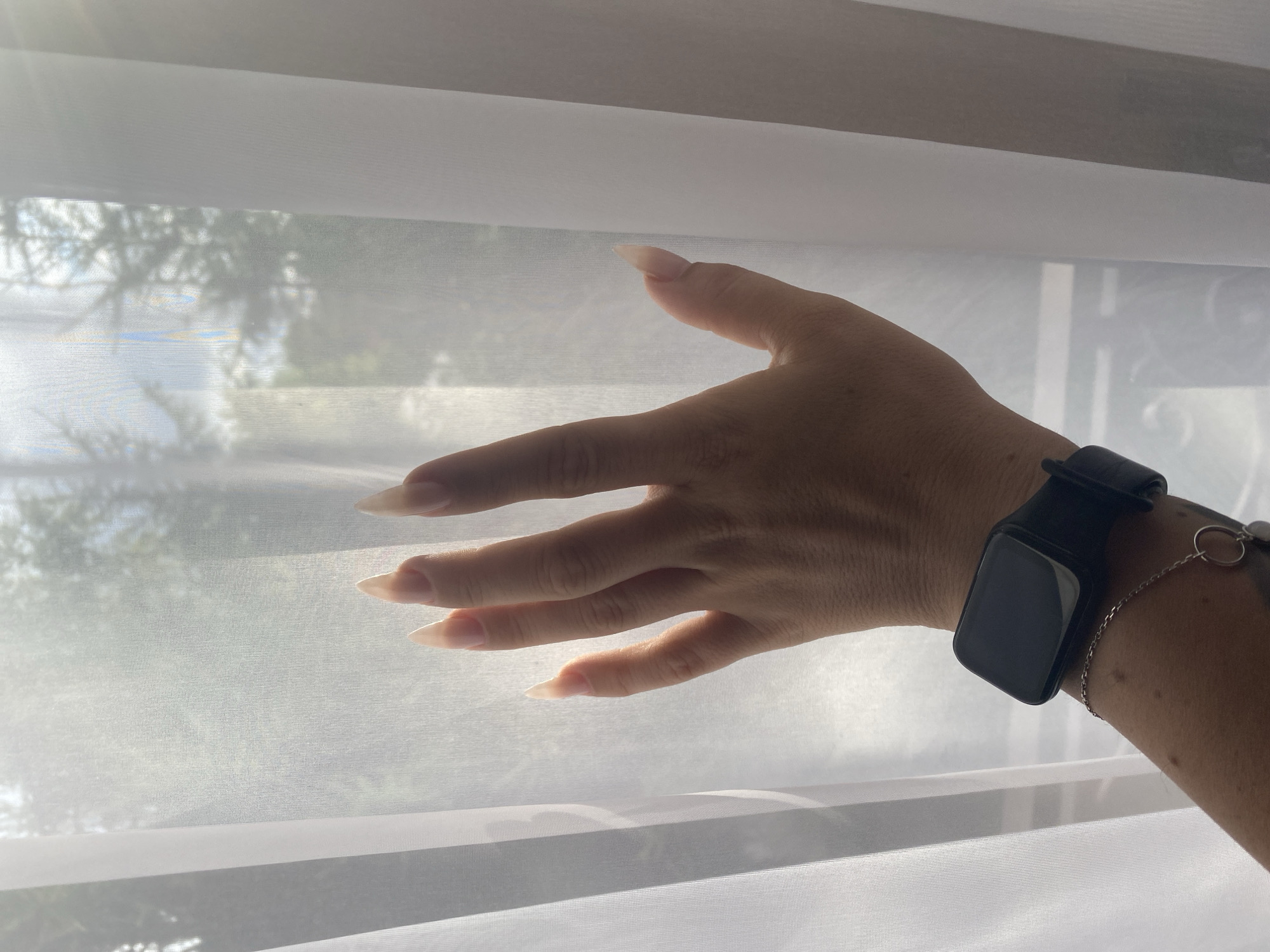 A close-up, vertical, eye-level shot focuses on a left hand with long, stiletto-shaped fingernails, illuminated by soft, diffused light filtering through sheer white curtains. The hand is positioned in front of the curtains, with the fingers slightly splayed and touching the fabric. The wrist is adorned with a black smartwatch and a delicate silver chain bracelet with a circular charm. Beyond the hand and curtains, the blurred background suggests an outdoor environment with trees and sky, indicating this scene is likely within a residential or building located near a natural setting. There is no specific information available about the location in the city of Krasnoflotskoe, Russia.