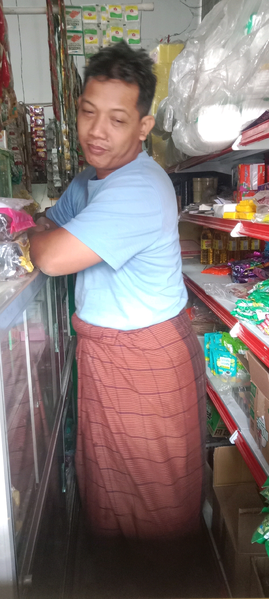 The image shows a man standing in what appears to be a small convenience store or a warung in Kras, Indonesia. The man is wearing a light blue t-shirt and a sarong that is red with thin black horizontal stripes. He has his arms crossed and is looking slightly downwards and to his left. The store shelves behind him are stocked with various items, including packaged goods, bottles of oil, and what appear to be snacks. The lighting suggests it is daytime, and the environment is enclosed. The man's expression is neutral. The overall impression is a candid shot within a retail setting.