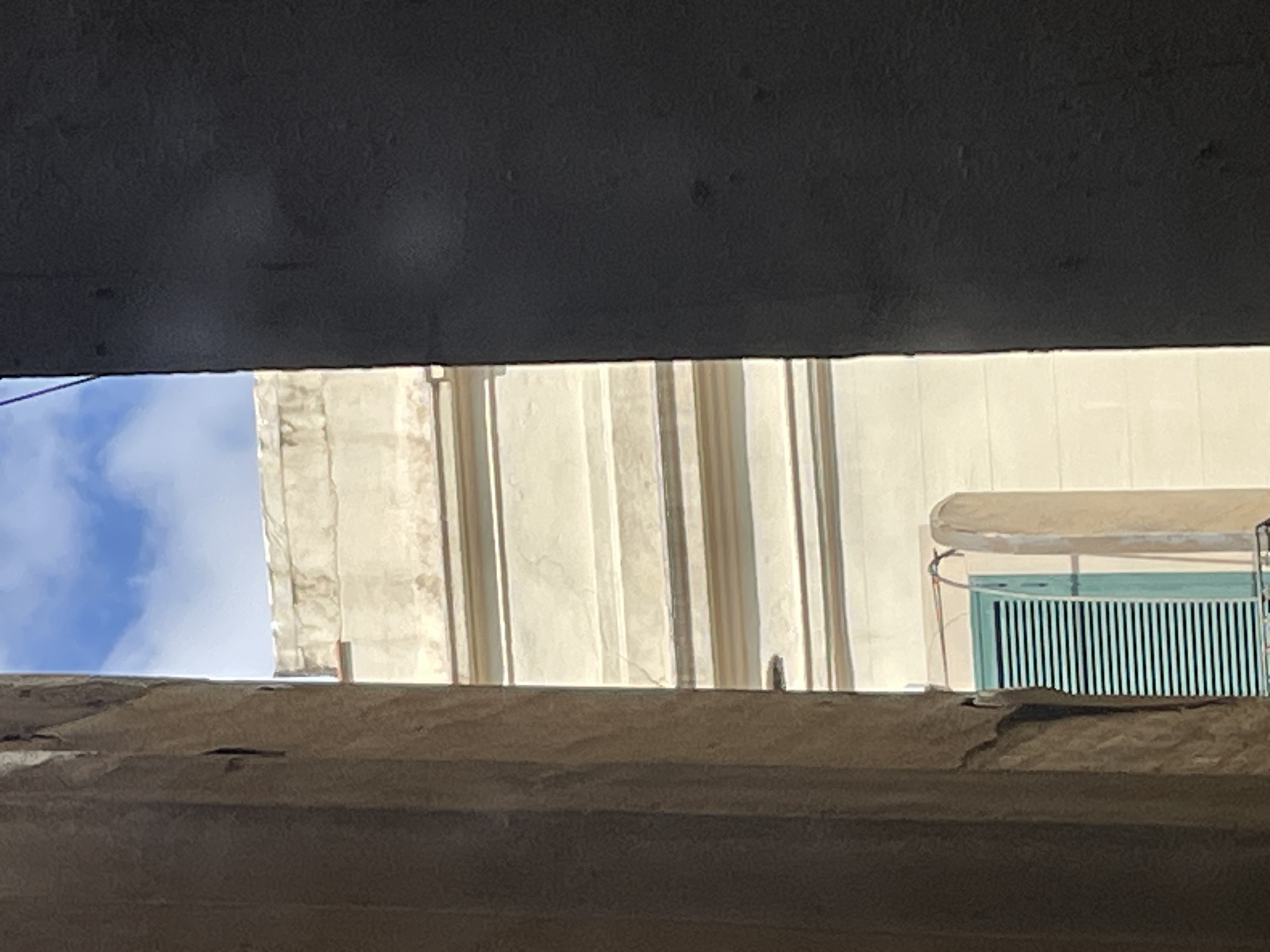 The image, taken from a low angle, possibly under an overpass or bridge, features a partial view of a building in Barcelona, Spain. The building is characterized by its cream-colored facade with vertical fluted architectural elements. A turquoise window with a metal grate is visible on the right side of the building. A glimpse of a blue sky with clouds can be seen on the left of the frame. The foreground is dominated by the underside of a concrete structure, obscuring the view.