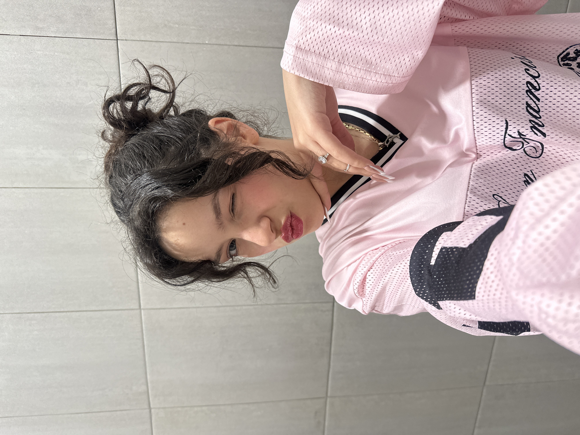 A young female individual is depicted in a chest-up portrait against a light grey tiled wall. Her dark, wavy hair is pulled into a messy bun, with loose strands around her face. She is winking with her right eye (viewer's left) and puckering her lips, which are covered in red lipstick. A small mole is visible on her forehead above her right eyebrow. She wears a light pink, long-sleeved jersey-style shirt with a mesh texture on the sleeves and lower body, and a smoother fabric panel on the upper chest. The V-neck collar features black and white stripes. Black lettering, partially visible as "Francisco," is present on the lower front of the shirt. Her right hand (viewer's left), adorned with a prominent silver-colored ring with a large clear stone on the ring finger and a smaller plain silver ring on the middle finger, is raised to her chin, with the index finger and thumb supporting her jawline. Her fingernails are long and manicured with white tips. A gold-colored chain necklace with a small pendant is visible at her neck. The background consists of evenly spaced, light grey rectangular wall tiles with visible grout lines. This scene is located in the city of Yerevan, Armenia.