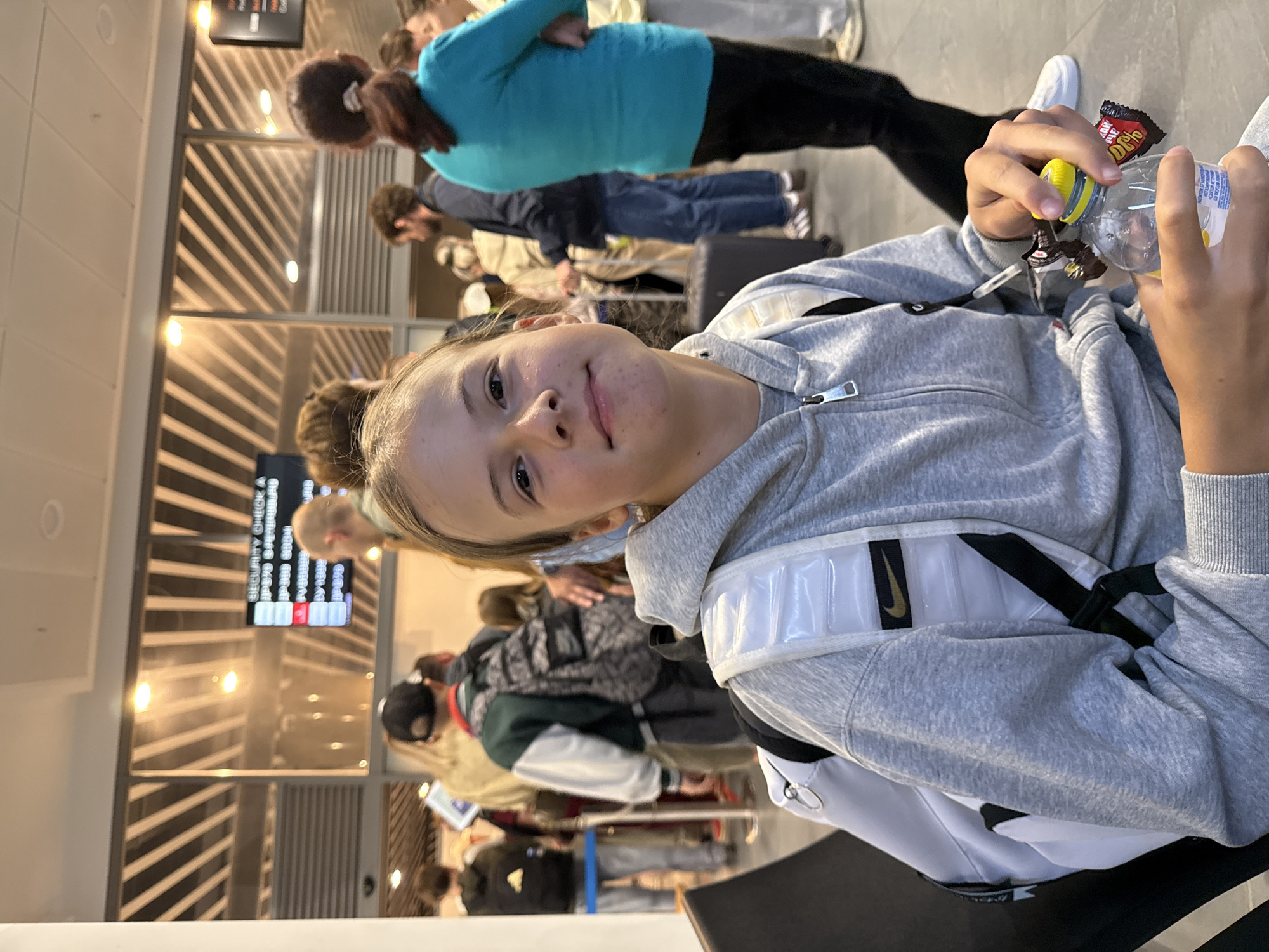 The image depicts a young person with blonde hair, wearing a grey hooded sweatshirt and a white backpack with black straps, holding a plastic bottle with a yellow cap and two candy bars. The person is seated in what appears to be an airport waiting area, with a security check sign visible in the background. Other people are also present in the background, some appearing to be in line. The lighting suggests an indoor environment, likely during daylight hours. The overall scene is casual and appears to be a moment of waiting. The text on the candy bars is partially visible and appears to be in Cyrillic. While the image is set in what appears to be an airport, specific details about the city of Bolshoye Savino, Russia, are not directly discernible from the visual content alone, though the presence of Cyrillic text on the candy could be a subtle indicator of the region.