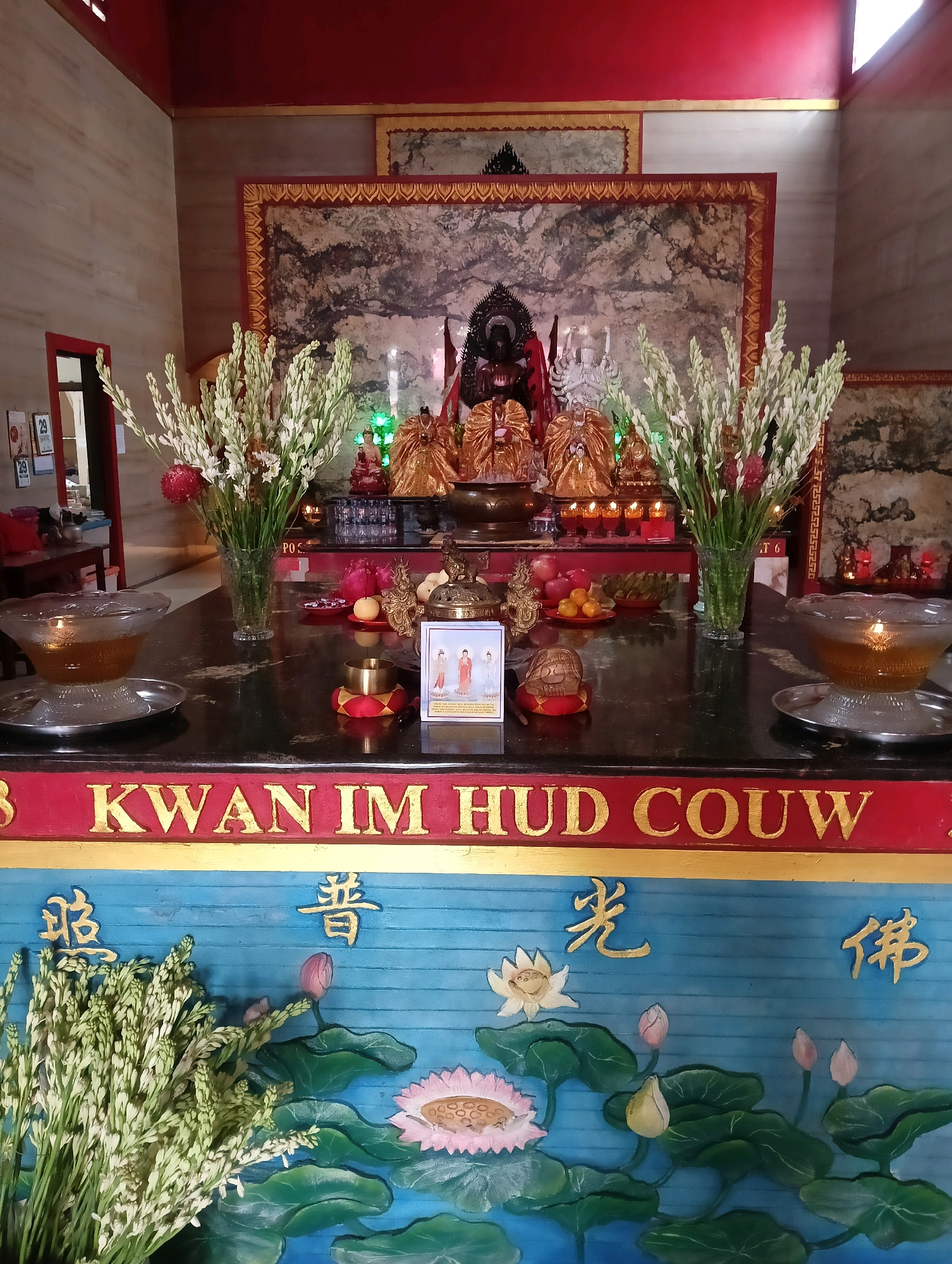 The image depicts an altar within a building, likely a temple or shrine, located in Rangkasbitung, Indonesia. The altar is adorned with various religious objects and decorative elements.
In the background, a large framed artwork, possibly a painting or tapestry, features a dark, mountainous landscape with streaks of white, suggesting mist or clouds. In front of this artwork, several golden statues of deities or revered figures are arranged. Some are seated, while others stand, all appearing to be in ceremonial attire. In front of the main statues, a small framed picture displays three figures in red and white robes.
The altar surface itself is dark and polished, reflecting the surrounding lights. On the left side of the altar, a clear glass bowl filled with a golden liquid, likely oil or tea for an offering, sits on a silver tray. To its right, a bouquet of white flowers in a glass vase is prominently displayed, with a single red flower among them. Further to the right, another clear glass bowl with a lit candle floats in a golden liquid is visible.
Scattered on the altar are several smaller objects, including what appear to be fruits (oranges and a pear-like fruit), small red objects, and a golden censer. A small brass bell rests on a red cushion.
A red and gold border runs across the front of the altar, with gold lettering that reads "KWAN IM HUD COUW." Below this, a blue panel features carved or painted lotus flowers in various stages of bloom, along with green lily pads. Golden Chinese characters are inscribed on this blue panel.
In the lower left corner of the image, a large bundle of the same white flowers seen in the vase on the altar is placed, suggesting a recent offering or preparation. The overall lighting suggests an indoor environment, possibly with natural light filtering in from the upper right, casting some shadows. The scene conveys a sense of reverence and tradition associated with religious practices.