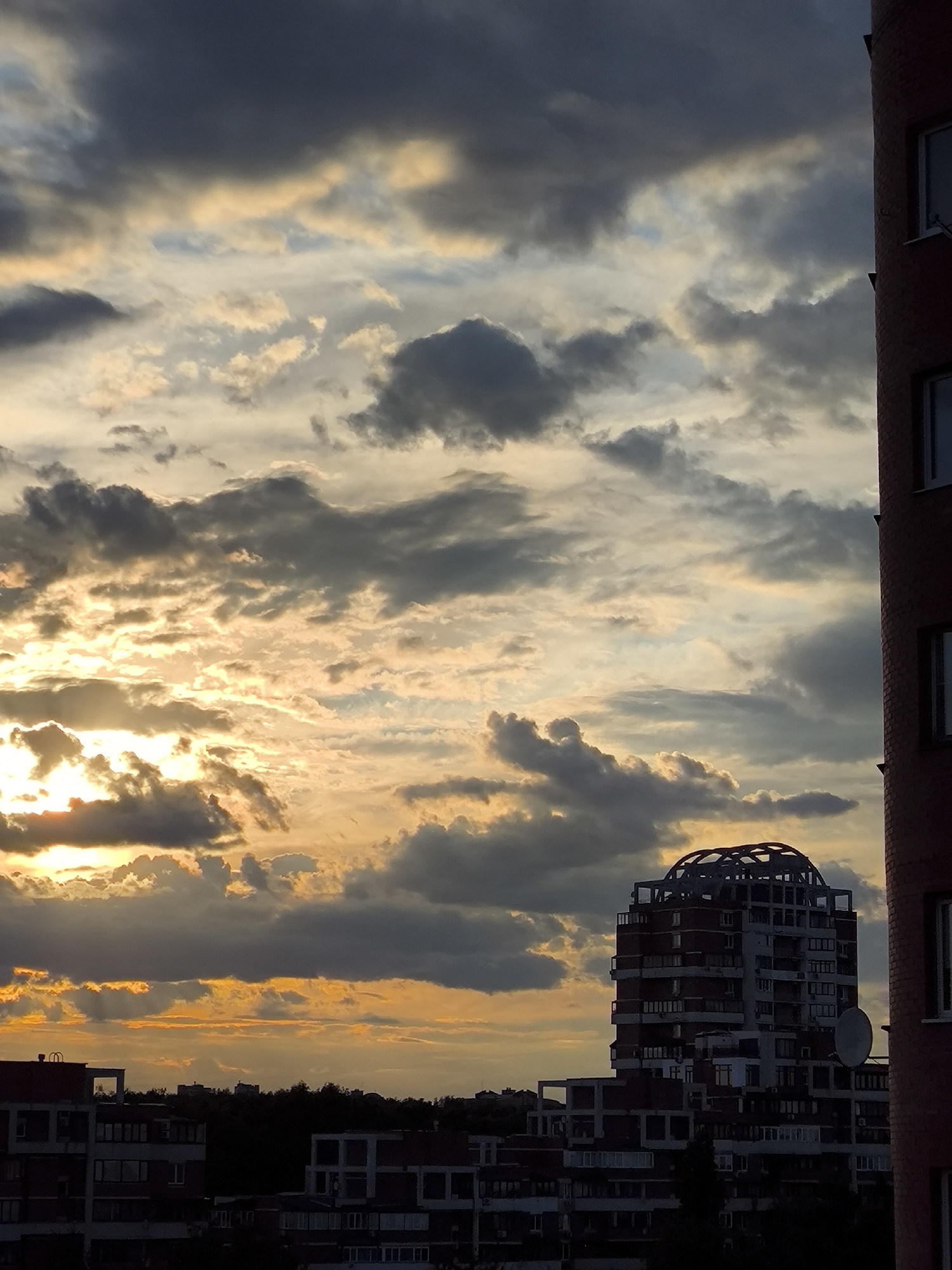 The image displays an urban or suburban cityscape captured during either sunrise or sunset, characterized by a dramatic and colorful sky. The sun, positioned low on the left, casts brilliant golden and orange light through gaps in the clouds, creating rays and illuminating the edges of some cloud formations. The sky is filled with a mixture of dark grey, lighter grey, and brightly lit, warm-toned clouds.
In the mid-ground, a distinctive, tall building with a unique architectural design stands out. It features a prominent, dome-shaped, open-work structure on its roof and multiple balconies and windows along its red and white facades. A large satellite dish is affixed to its side, clearly visible against the sky. To the far right, a section of another reddish-brown brick building with white-framed windows is partially visible, suggesting it's very close to the viewer's perspective. Several other blocky residential buildings, appearing mostly in silhouette, are scattered across the lower left and central background, with a dark treeline or distant structures forming the horizon.
No people or discernible activities are visible in the frame. The time of day is clearly either early morning or late evening, as indicated by the low sun and the warm, intense lighting. The overall mood conveyed is one of natural beauty and urban grandeur during a transitional period of the day. There is no visible text in the image.