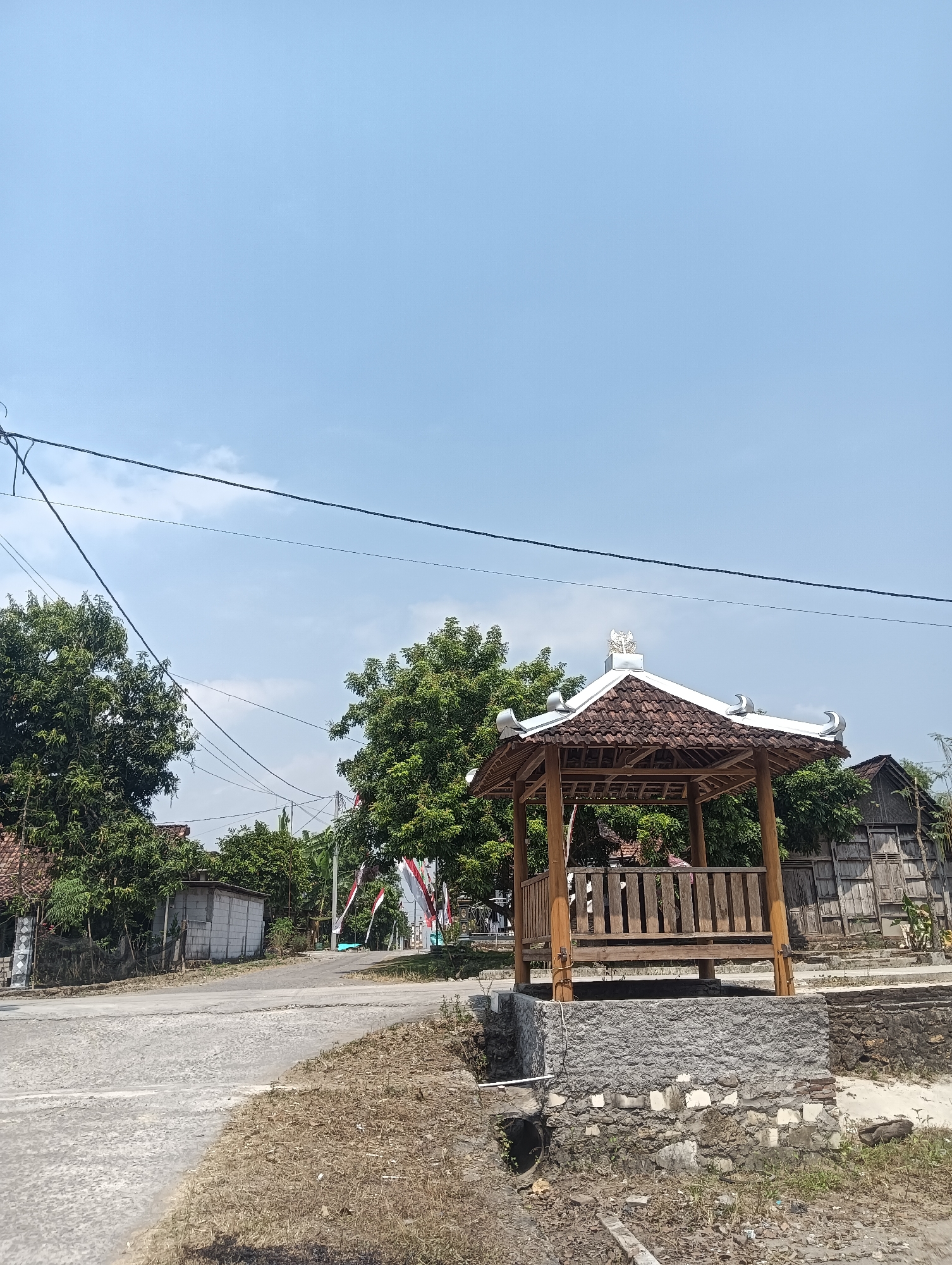 This image depicts an outdoor scene in the city of Tanggan, Indonesia, under a clear blue sky with a few scattered clouds. In the foreground, a paved road curves to the left, bordered by dry grass and vegetation. To the right of the road, a small, traditional wooden gazebo with a tiled roof stands on a stone foundation. The gazebo has wooden railings and a bench inside. Behind the gazebo, a weathered wooden building with a textured facade is visible.
Further back, lush green trees flank the scene, with some houses and smaller buildings nestled amongst them. Several Indonesian flags, displaying red and white stripes, are fluttering on poles, suggesting a possible celebration or national event. Power lines crisscross the sky, indicating the presence of infrastructure. The overall impression is of a quiet, rural or semi-rural setting in Indonesia on a bright day.