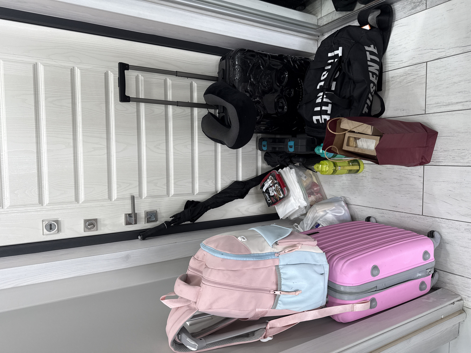 The image shows a collection of luggage and personal items on what appears to be a shelf or ledge. The items include two suitcases (one black, one pink), two backpacks (one black, one pink and blue), a neck pillow, an umbrella, a water bottle, and other miscellaneous items in plastic bags and containers. The background includes a white paneled door and what looks like a tiled wall. There is no explicit indication that the scene is in Sochi, Russia, or any visible people.
