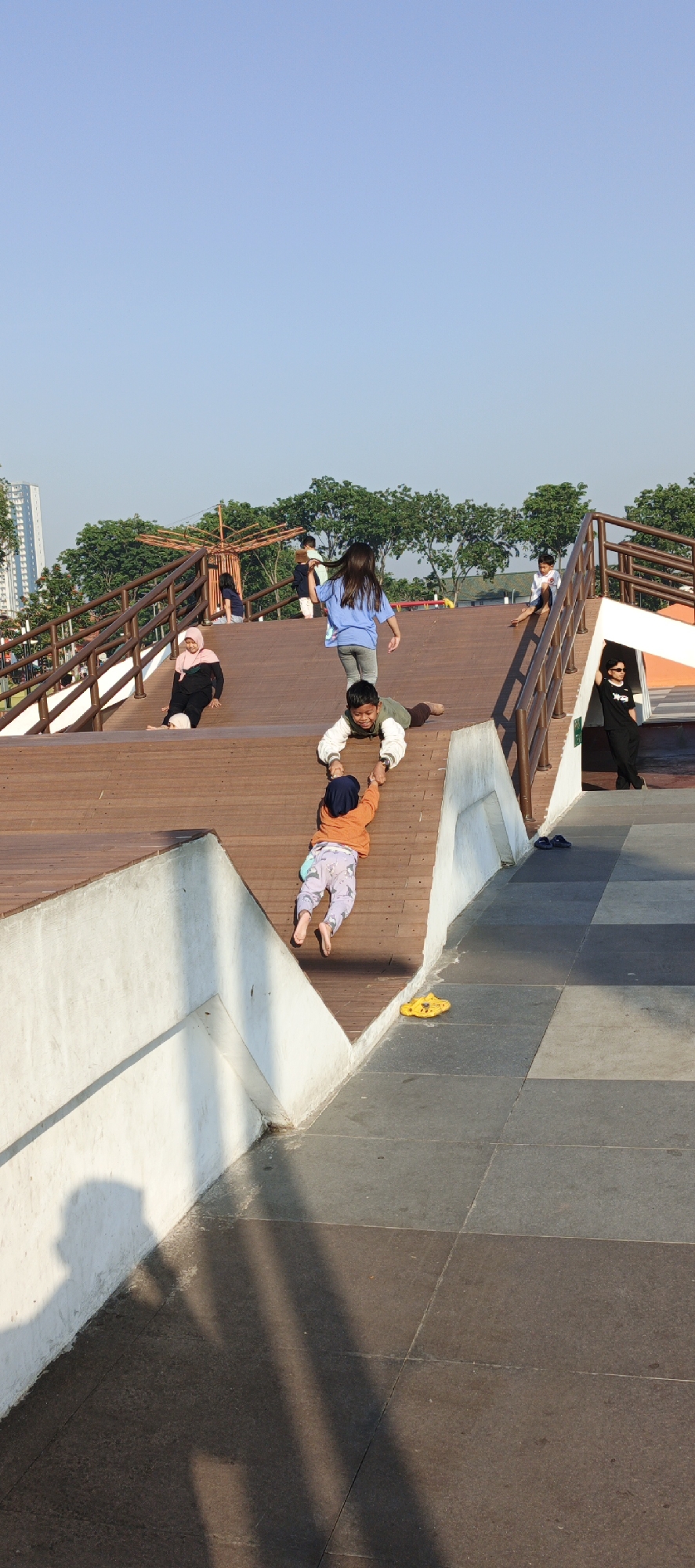 The image depicts a bright, sunny day in Sidoarjo, Indonesia, likely at a park or recreational area. In the foreground, two children are interacting on a wooden ramp-like structure. The older child, wearing a green and white jacket and brown pants, is lying on their stomach, holding the hands of a younger child. The younger child, wearing an orange long-sleeved shirt, blue headscarf, and patterned pants, is being pulled up by the older child. Their feet are bare. A bright yellow sandal is visible on the tiled ground to the right.
In the middle ground, a girl in a blue t-shirt and grey pants is walking down the ramp. Further up the ramp, a woman wearing a pink hijab and black attire is sitting down. Other children and individuals are visible in the background, some walking or sitting on the wooden structure and stairs. The structure itself features wooden planks and railings, suggesting it's part of a playground or elevated walkway. Trees and a tall building are visible in the distant background under a clear blue sky. The scene suggests a casual, recreational setting where families are enjoying outdoor activities.