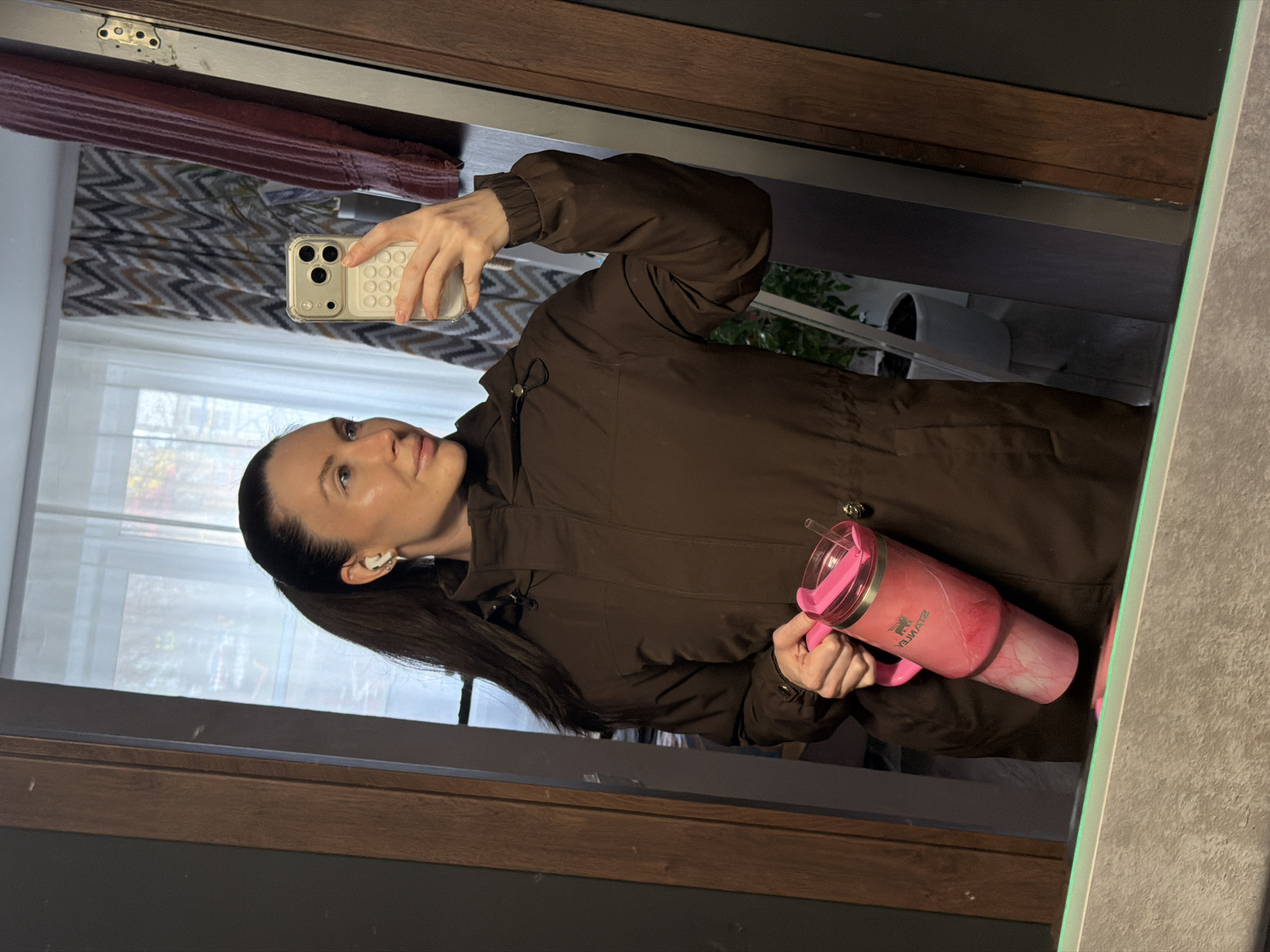 The image displays a mirror selfie of a woman taken indoors, rotated 90 degrees counter-clockwise. The woman, with long dark hair styled in a ponytail, wears a dark brown jacket or coat and white wireless earbuds. She holds a light-colored smartphone with a textured or "pop-it" style case in her right hand, using it to capture her reflection. In her left hand, she holds a large, pink and white marbled tumbler with a handle and a clear straw. The brand name "STANLEY" is visible in reverse on the tumbler, along with what appears to be "PLAST" (also reversed) near the bottom.
The background visible in the mirror's reflection includes a window with sheer white curtains, suggesting natural daylight. Outside the window, a blurred view hints at trees or buildings, indicating an urban or suburban setting. Below the window, a patterned fabric with a grey and brown chevron or zigzag design is discernible. Wooden and dark-colored furniture or architectural elements frame parts of the mirror. The woman has a composed, neutral expression, gazing slightly upwards. The scene takes place during the daytime in Perm, Russia.