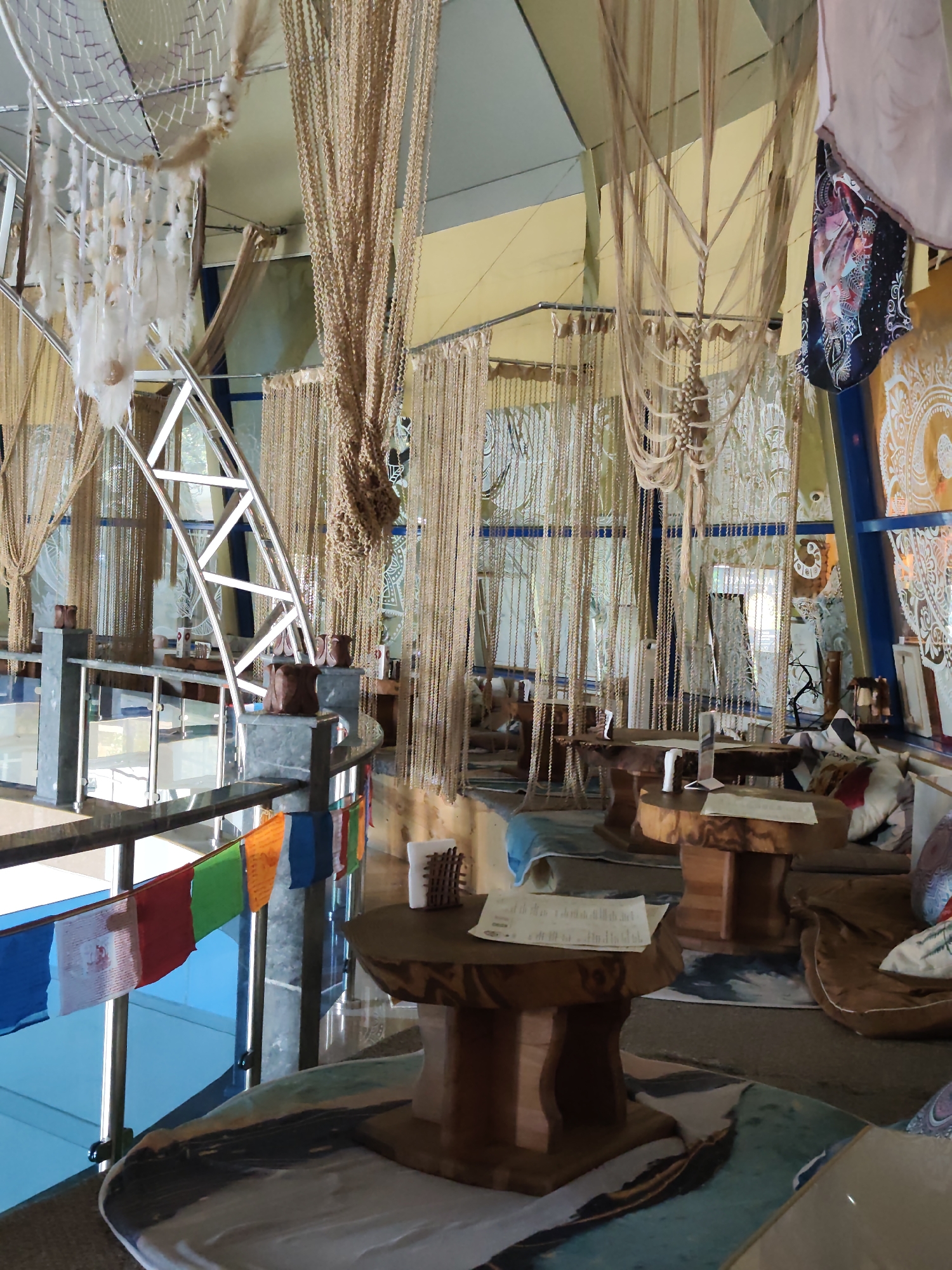 The image depicts the interior of a cafe or restaurant, possibly located in Sochi, Russia, given the Tibetan prayer flags visible. The decor is eclectic and bohemian, featuring numerous hanging macrame and beaded curtains, dreamcatchers, and various fabric drapes. Natural wood elements are prominent in the furniture, with several rustic wooden tables and stools. The seating area appears to be arranged around these tables, with comfortable floor cushions visible. The lighting is diffused, suggesting an indoor space. The background reveals large windows or glass partitions, allowing some natural light to filter in. The overall impression is one of a relaxed, spiritual, or artistic ambiance. There are no people visible in the image.