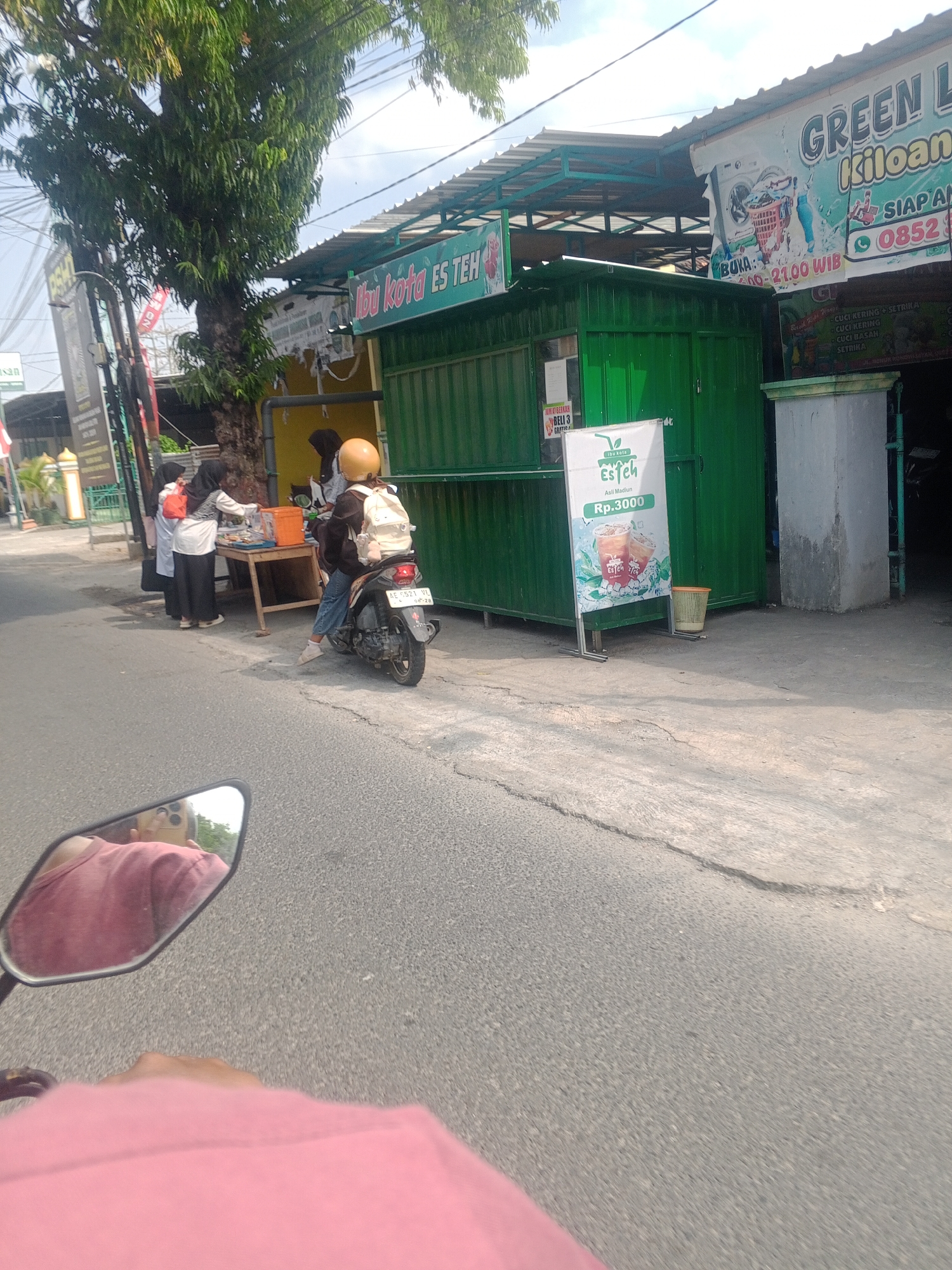 In Ronowijayan, Indonesia, a street scene unfolds in front of a green food stall labeled "Ibu Kota Es Teh." A person on a scooter, partially visible in the foreground, is reflecting in the scooter's mirror, showing the user holding the phone and wearing a pink shirt. The scooter is parked near the stall. To the left of the stall, two individuals stand at a table where drinks are being prepared, with one person wearing a headscarf and another in a white shirt. Further left, a large tree shades the area, and a sign for a laundry service with the name "GREEN LAUNDRY" is visible, displaying "KILOAN" and a phone number. The pavement in front of the stall is cracked. The sky is overcast.
