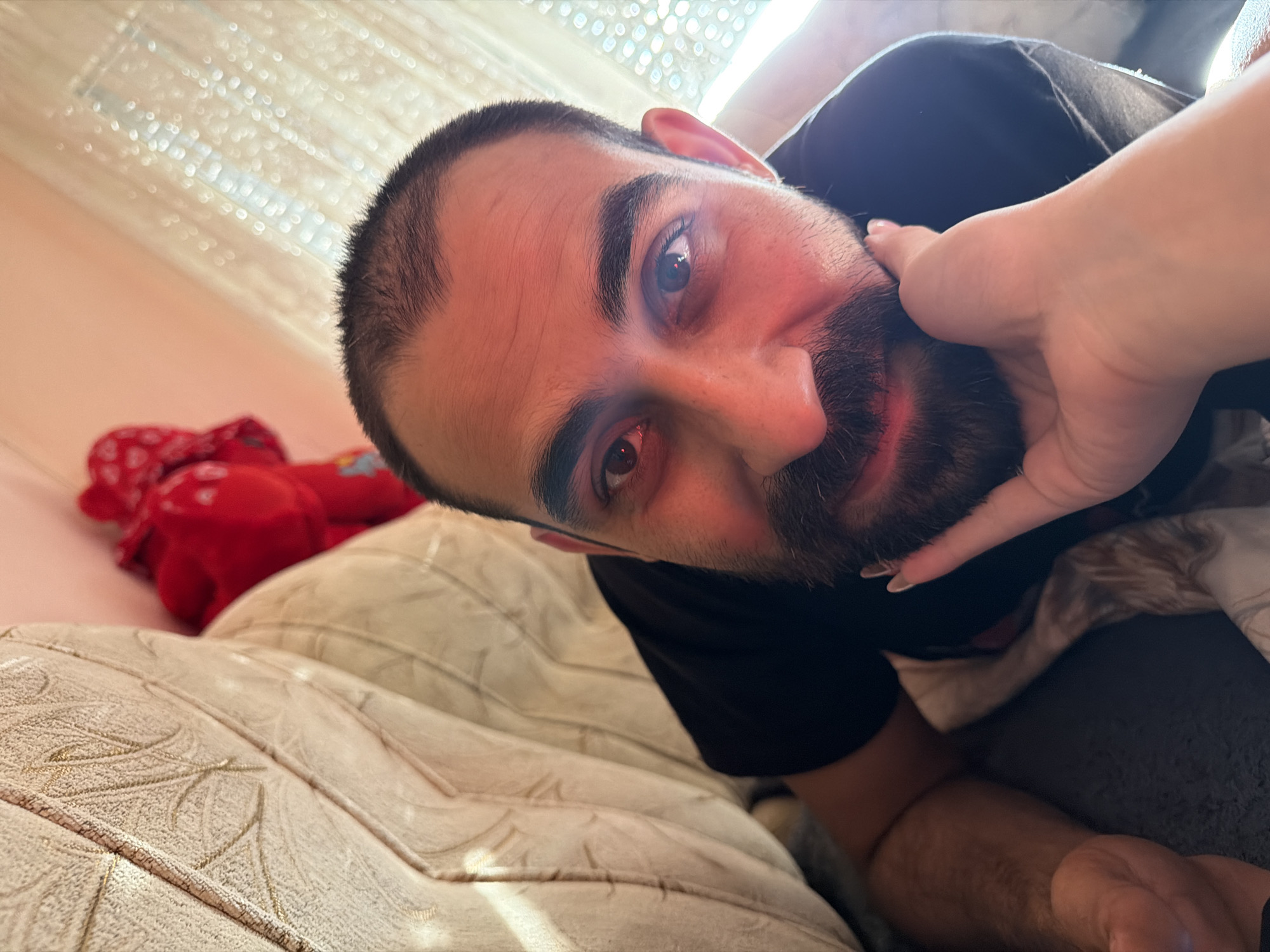 This close-up, Dutch-angle shot captures a man lying down, looking directly at the camera. His head is tilted, with his right ear and part of his forehead visible. He has dark, short hair, thick eyebrows, and a full beard. His eyes are dark and appear to be reflecting light. A woman's hand is gently touching his cheek and jawline.
The man is wearing a black t-shirt. He is lying on a beige, tufted surface, possibly a sofa or bed, with a textured pattern. In the background, to the left, a red plush toy with a white polka-dot pattern is visible, partially obscured by the man's head. The overall lighting suggests daytime, with bright light illuminating the scene from above and to the right. The setting appears to be indoors, possibly a bedroom or living room, with a hint of a window or curtain in the upper background. There is no visible text in the image. The interaction between the man and the hand suggests a moment of intimacy or affection. The location is Stara Zagora, Bulgaria.