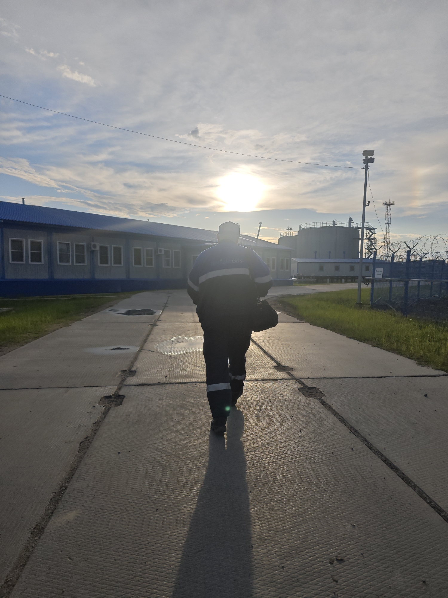 The image depicts a solitary individual walking away from the camera on a concrete path in an industrial or semi-industrial area in Unknown, Russia. The person, dressed in blue and dark blue workwear with a white reflective stripe on the arm and carrying a dark bag over their left shoulder, casts a long shadow extending towards the viewer due to the low sun. The sun is positioned directly behind the person, causing significant backlighting and a lens flare effect.
To the left of the path, a long, single-story building with a blue corrugated metal roof and light blue walls features numerous white-framed windows. To the right, a grassy strip runs alongside a fence topped with barbed wire. Beyond the fence, various industrial structures are visible, including large, cylindrical storage tanks and a tall, lattice communication or observation tower. The sky is partly cloudy, displaying light blue hues and scattered white clouds. A faint parhelion (sun dog) is present to the right of the sun. The concrete path consists of textured rectangular slabs with visible cracks and minor debris.
