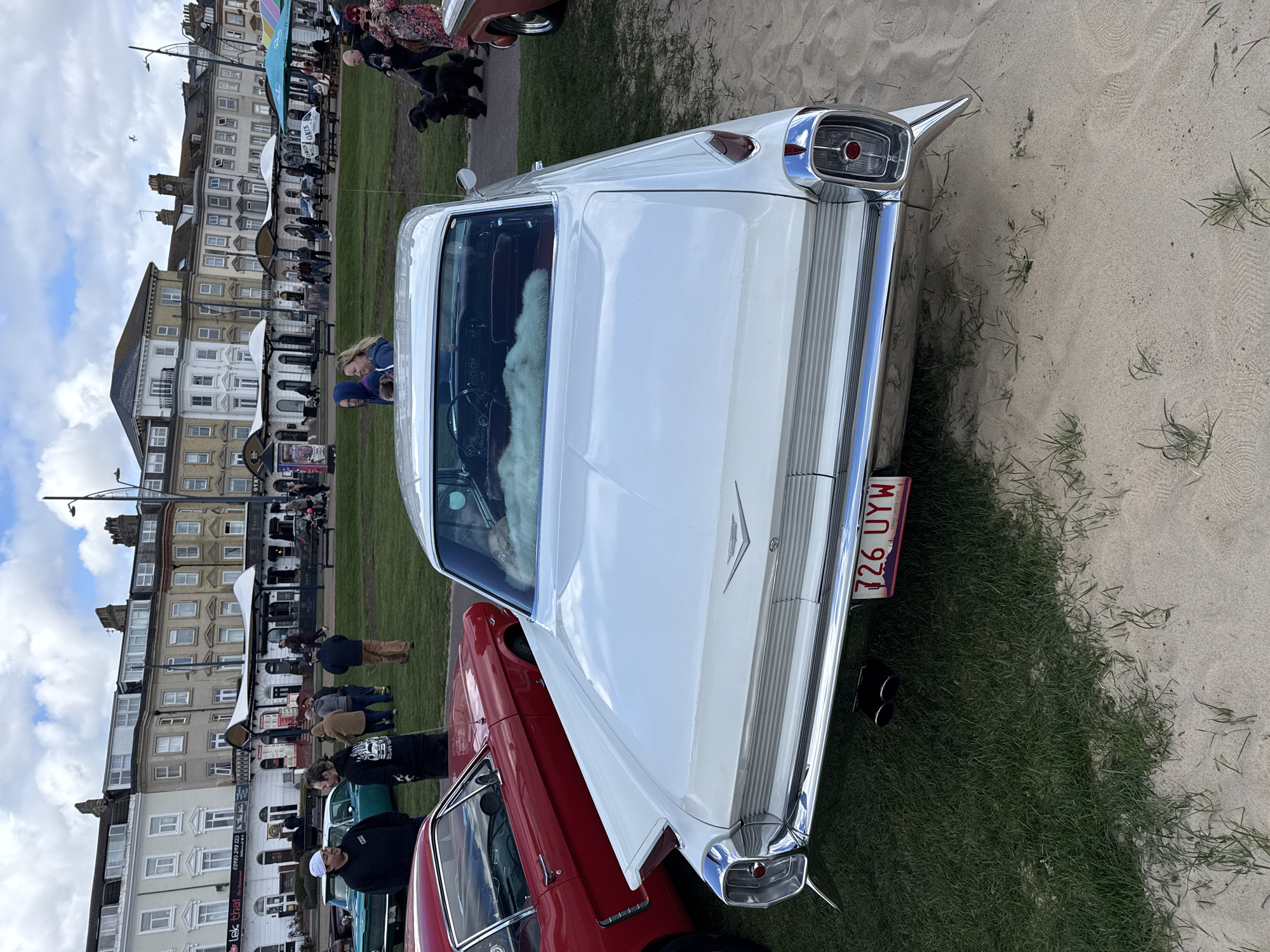 The image captures the rear view of a white vintage car, possibly a Cadillac, parked at a car show held along the promenade in Great Yarmouth, United Kingdom. The car features prominent tailfins and chrome trim, and its license plate reads "726 UYW." The car is parked on a grassy area adjacent to the sandy beach. To the left of the white car is part of a red car. In the background are several buildings, including shops and apartments. Several people are milling around, some of them looking at other cars. The sky is cloudy with a few patches of blue.
