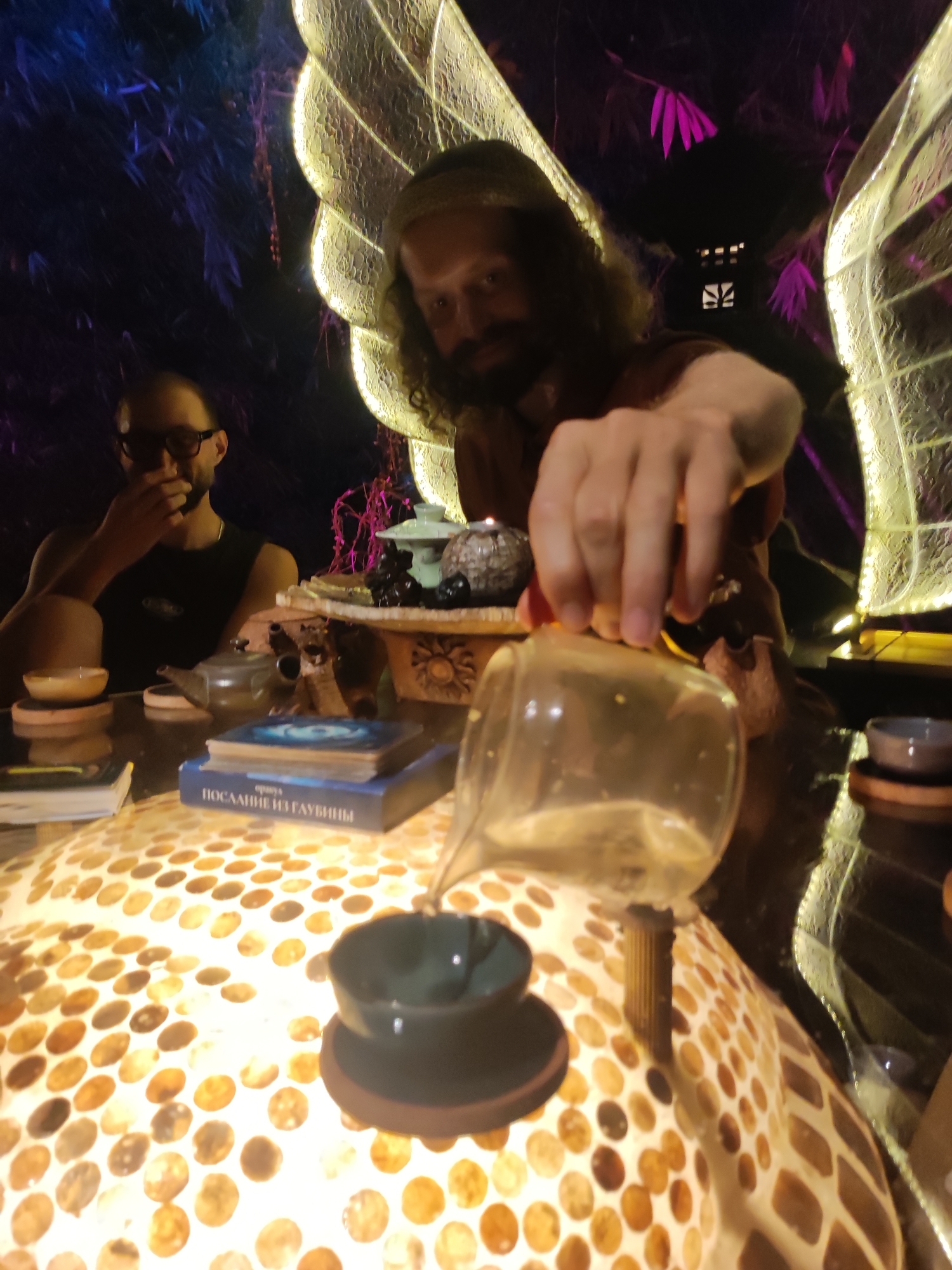 In a dimly lit interior in Lokaserena, Indonesia, a bearded man with long hair is pouring liquid from a glass pitcher into a small teacup set on a round, mosaic-tiled table. Another man with glasses sits in the background, partially obscured. The table holds various objects including books ("ПОСЛАНИЕ ИЗ ГЛУБИНЫ"), teapots, and small bowls. Illuminated wings, possibly part of the decor, are visible behind the pouring man. The scene appears to be a tea ceremony or gathering.