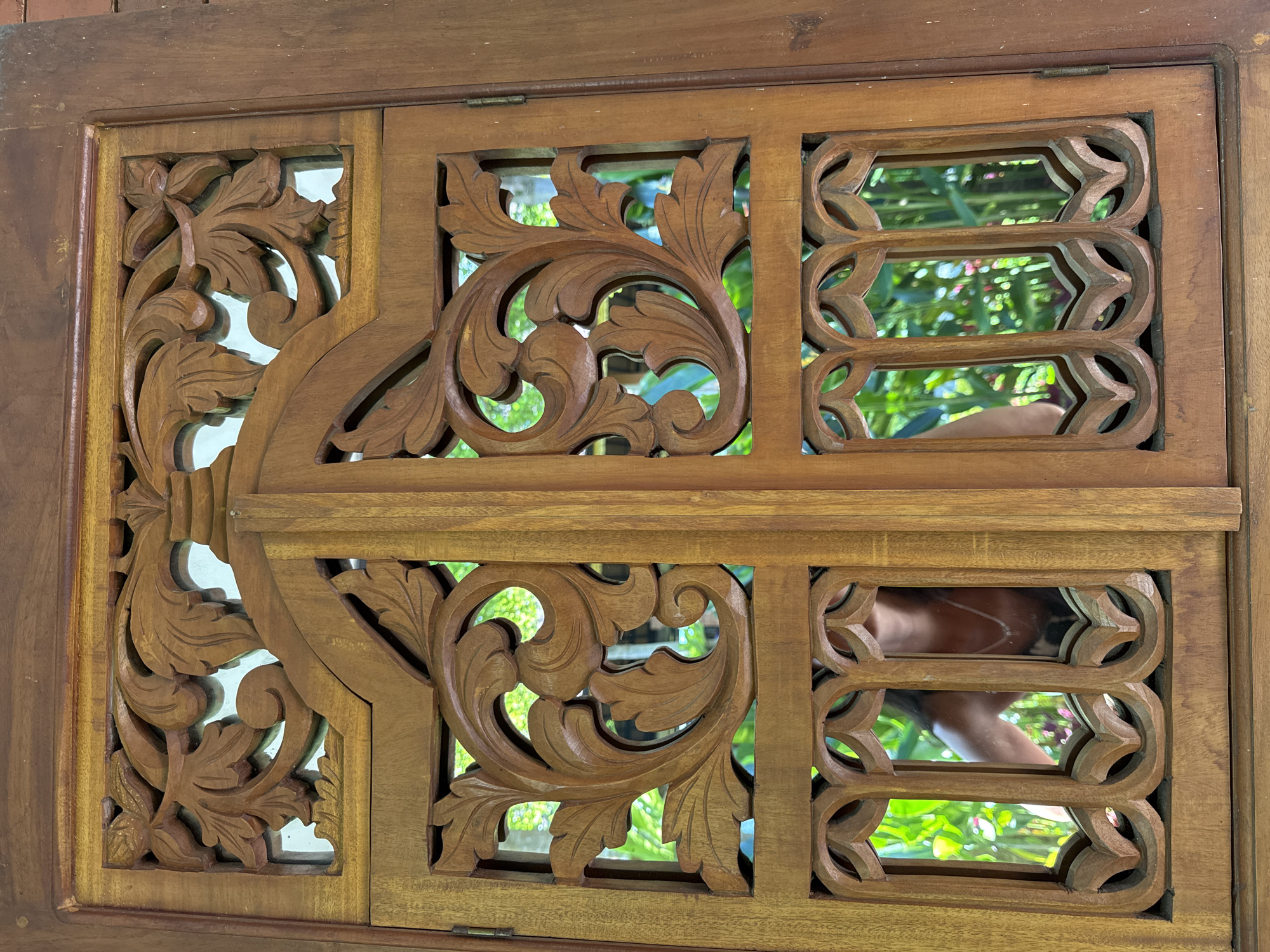 The image depicts a detailed wooden carving, likely part of a door or window frame, consisting of three distinct rectangular panels. The panel on the left showcases intricate floral designs carved in wood. The middle panel also features leaf carvings, but with larger open spaces. The right panel displays a repeating geometric design with smaller openings. The background visible through the carvings is blurred, suggesting an outdoor scene with greenery. Based on the location in Rendang, Indonesia, it can be inferred that the wood is probably of Indonesian origin. In the reflection on the right panel, an arm with a silver necklace or chain are visible indicating the presence of a person.