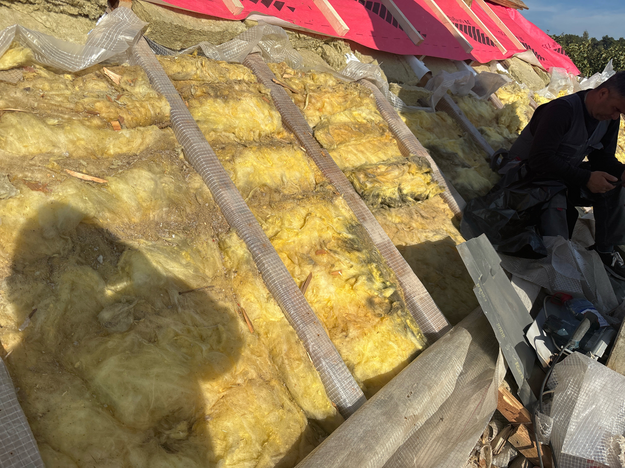 The image depicts the insulation of a rooftop in Zhavoronki, Russia. The insulation material, primarily yellow fiberglass, is exposed and appears to be in the process of installation or repair. Several wooden beams are visible, supporting the structure. Translucent plastic sheeting, possibly a vapor barrier, is draped over some sections of the insulation. In the background, pink roofing material and wooden framing are present, indicating ongoing construction. A person, dressed in work attire including a vest and pants, is seated on the right side of the image, looking down at a device, likely a mobile phone. The environment suggests an outdoor setting on a sunny day.