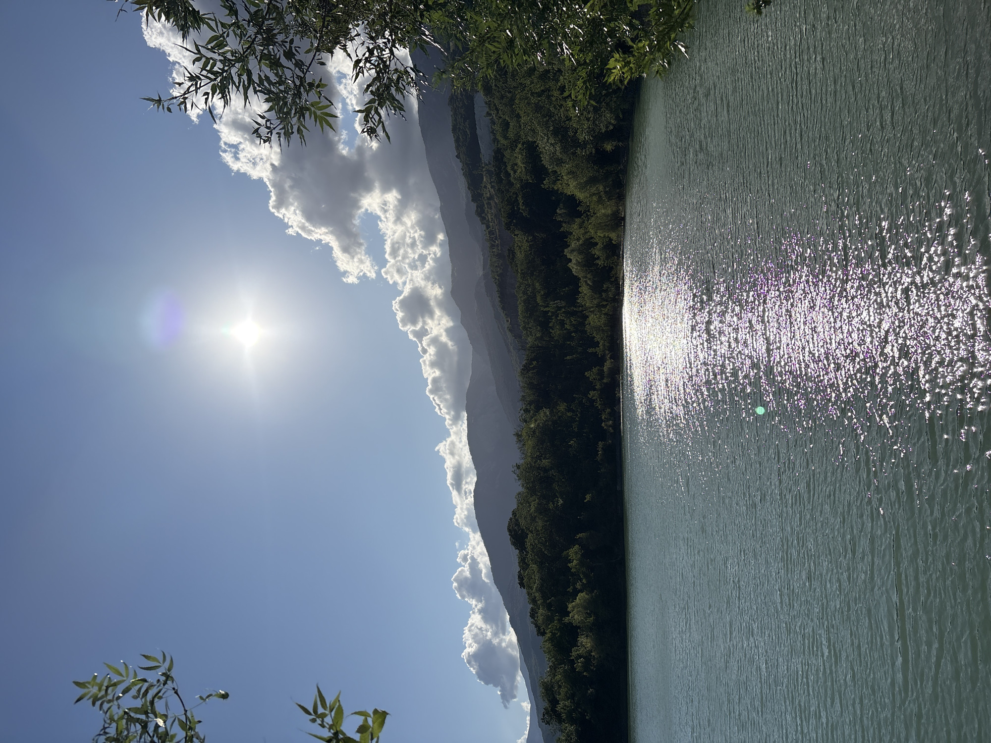 The image depicts a tranquil lake scene with a backdrop of lush green hills and a bright sky. In the foreground, the calm water of the lake reflects the sunlight, creating a shimmering effect. The middle ground features a dense forest covering the slopes of the hills. The background is dominated by majestic mountains, partially obscured by fluffy white clouds. The sun is shining brightly in the sky, casting a warm glow over the landscape. The image appears to have been taken in the city of Kiparisnoe, Ukraine, given the characteristic natural beauty of the region. No people or specific objects are visible in the scene, emphasizing the natural serenity of the environment.
