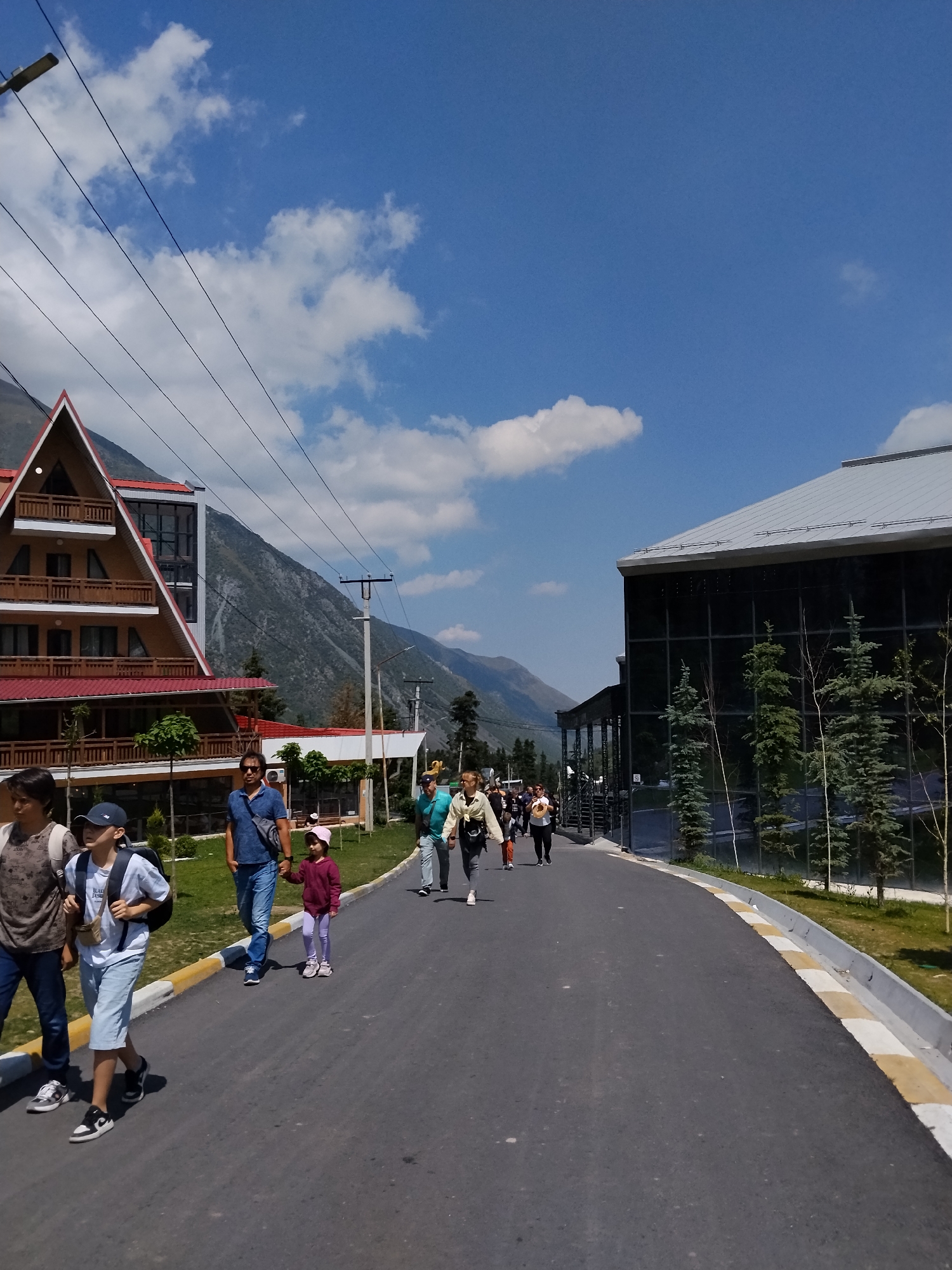 In this outdoor shot taken on a sunny day in an unknown city in Kyrgyzstan, a group of people are walking on an asphalt road. The road is bordered by a strip of grass and a curb with yellow and white markings.
To the left, a distinctive A-frame building with a wooden facade, red roof, and multiple balconies dominates the foreground. Behind it, a steep, rocky mountain slopes upwards. Above and behind the building, power lines are visible against a blue sky with scattered white clouds.
To the right, a modern building with a dark glass facade and a grey, sloped roof stands out. Several evergreen trees are planted in front of this building.
The people in the image are walking in the same direction. In the foreground, a young boy with a backpack, wearing a light blue t-shirt and shorts, walks alongside a woman in a brown shirt. Further ahead, a man in a blue polo shirt and jeans walks hand-in-hand with a young girl in a purple sweater and pink hat. A group of individuals is also seen walking further down the road, some of whom are wearing light-colored clothing. The overall atmosphere appears to be one of casual exploration or travel.
