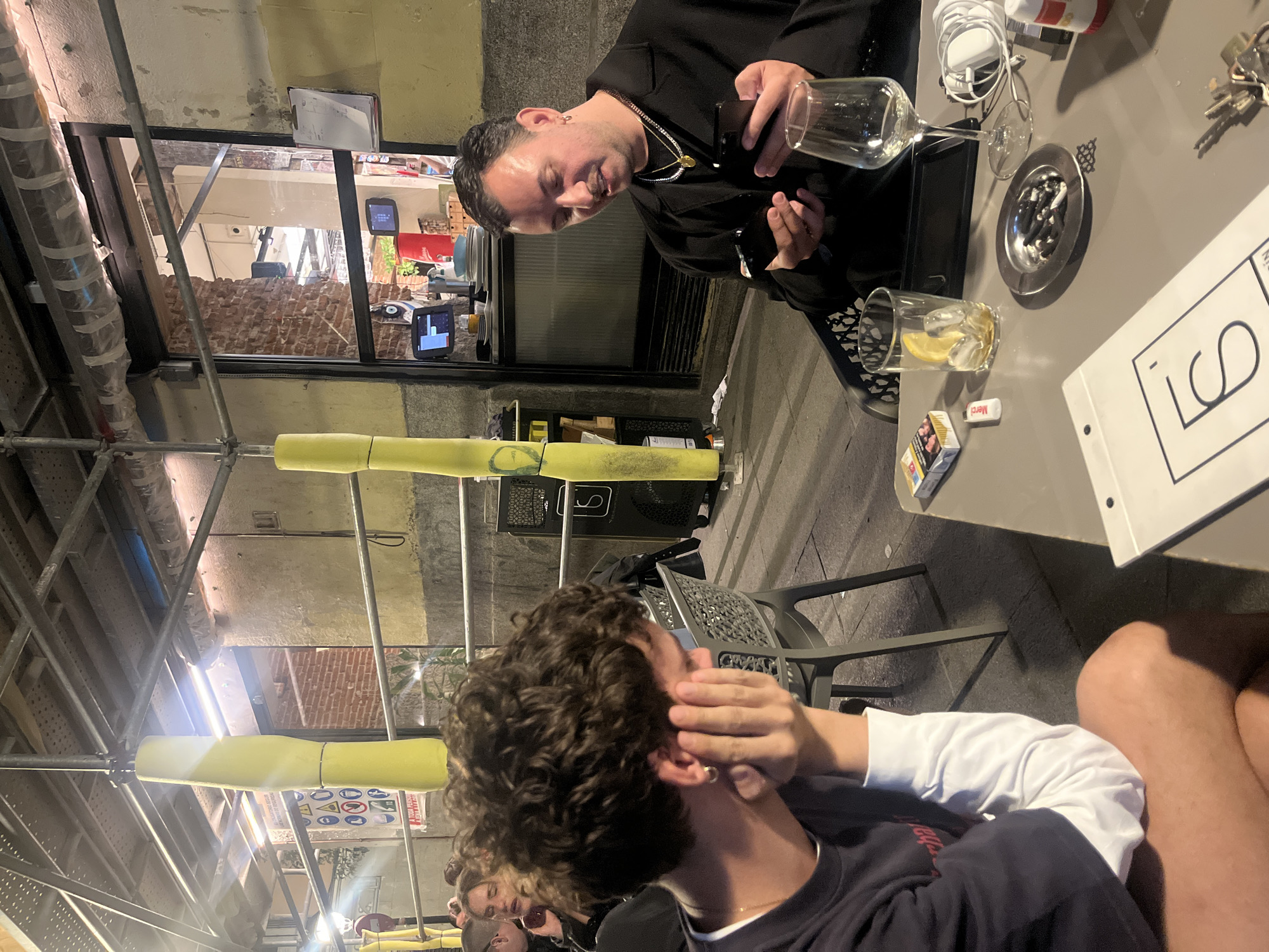 The image depicts an outdoor seating area, possibly a restaurant or bar, with scaffolding visible overhead, suggesting ongoing construction or renovation. One man, dressed in a black shirt and gold chain, is seated at a table, looking down at a cell phone in his hands. The table contains several items: a wine glass, a glass with a lemon slice, cigarettes, an ashtray with cigarette butts, keys, a charger, and a paper menu. Another person, with curly brown hair, is seated at a table in the foreground. The person's hand is raised towards their face. Additional individuals are visible in the background. Based on the architectural style and general ambiance, the image is likely taken in Madrid, Spain.