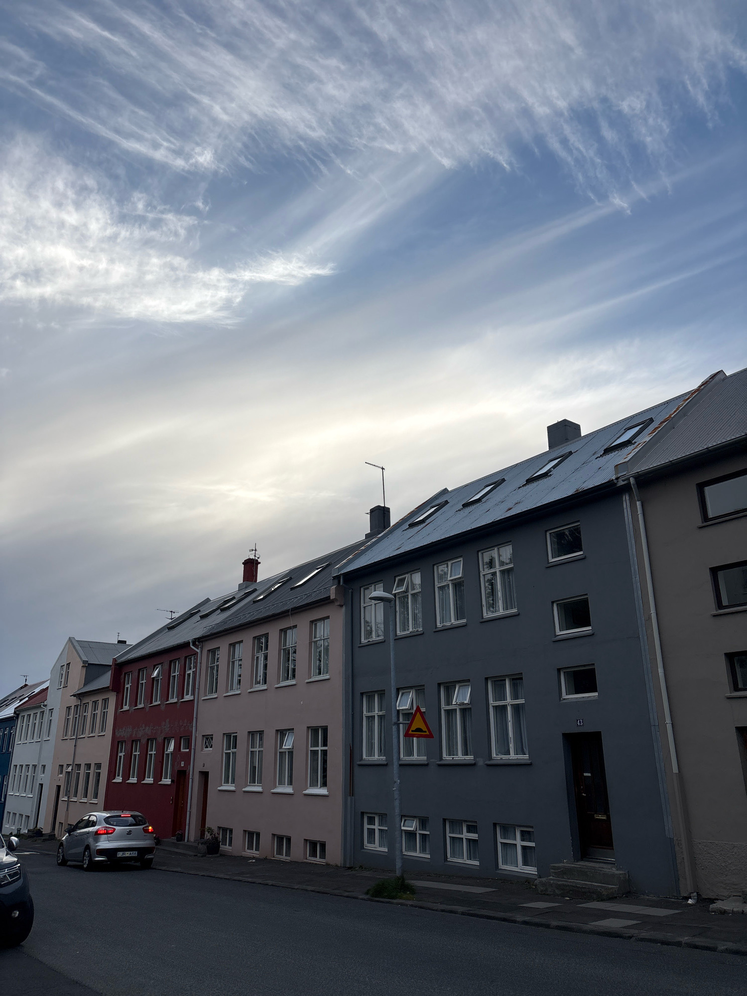 A row of colorful buildings lines a street in Reykjavik, Iceland, under a sky filled with wispy, elongated clouds. The buildings, in shades of pink, red, gray, and beige, have multiple windows and some have dormer windows on their sloped roofs. A silver car is parked on the side of the street, and another dark-colored car is partially visible in the foreground. A yellow traffic sign with a black symbol of speed bumps is posted on a pole in front of one of the buildings. The overall scene suggests a quiet urban environment, possibly in the early morning or late evening due to the lighting conditions and the dramatic sky.