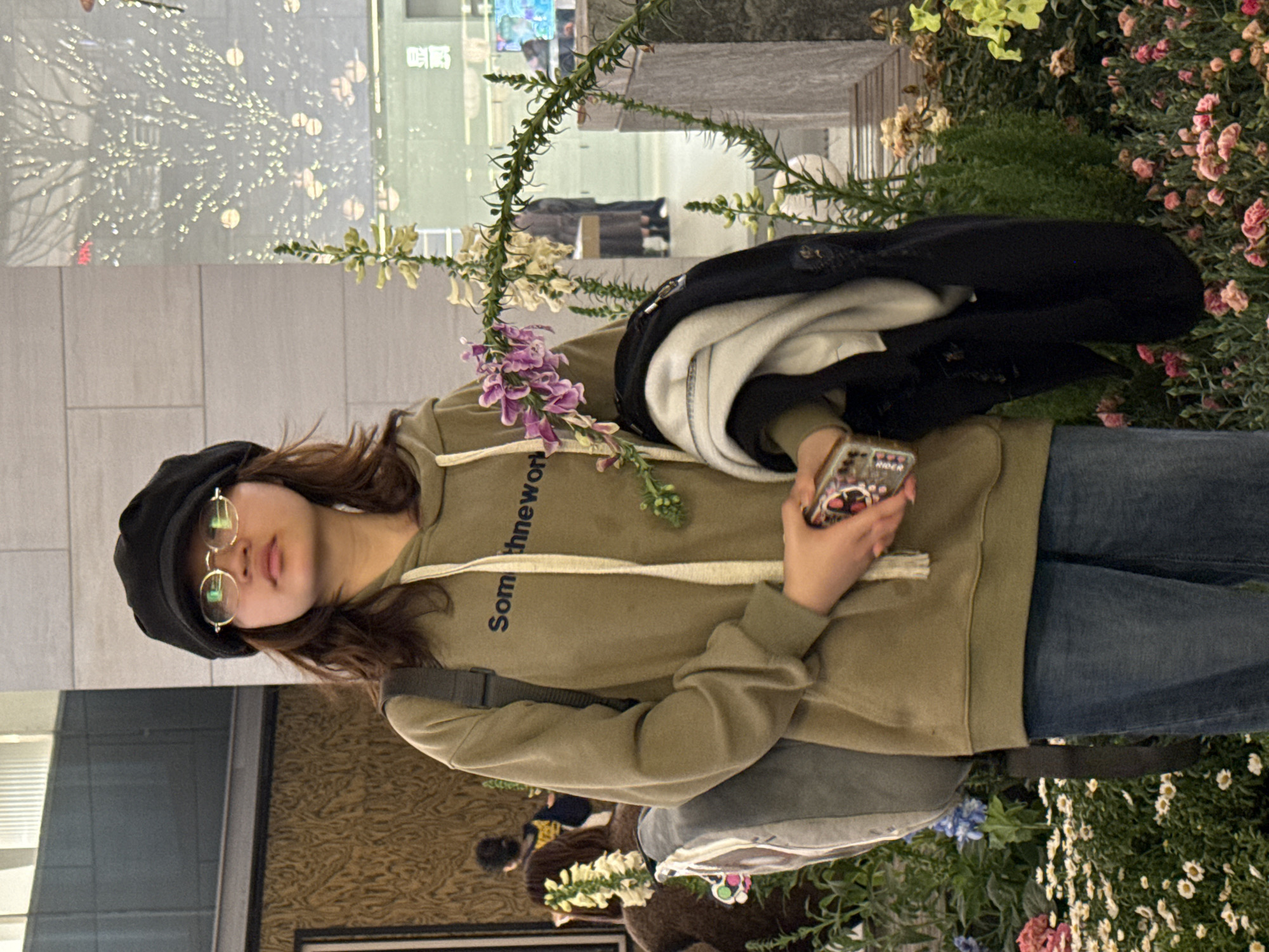 An East Asian female stands centrally within a brightly lit, modern indoor public space in Liangjiang, China, characterized by extensive floral displays. The subject wears a black newsboy cap, round gold-rimmed glasses, an olive green hooded sweatshirt with visible white drawstrings and partial text "Som thnewor" on the chest, and blue jeans. Her long, wavy brown hair extends from beneath the cap. A grey backpack is slung over her left shoulder. She holds a smartphone with a colorful patterned case in her right hand, while a black jacket and a lighter-colored garment are draped over her left arm. She is looking slightly upwards and to her right.
The immediate foreground and surrounding area are adorned with various live plants and flowers, including prominent tall stalks of purple and white bell-shaped blooms resembling foxgloves. Clusters of small white daisy-like flowers, pink carnation-like flowers, and blue blossoms are visible at ground level, interspersed with green foliage. The background features modern architectural elements: light-colored tiled walls, dark wooden or paneled surfaces, and large glass panels. These glass panels reflect diffuse lights, appearing as a tree-like pattern, and blurred distant figures. Chinese characters "显贝" are discernible in the reflection on a glass panel to the right. A blurred figure with dark hair is partially visible to the left behind the subject, and two additional blurred figures are seen in the distance through the glass reflection.