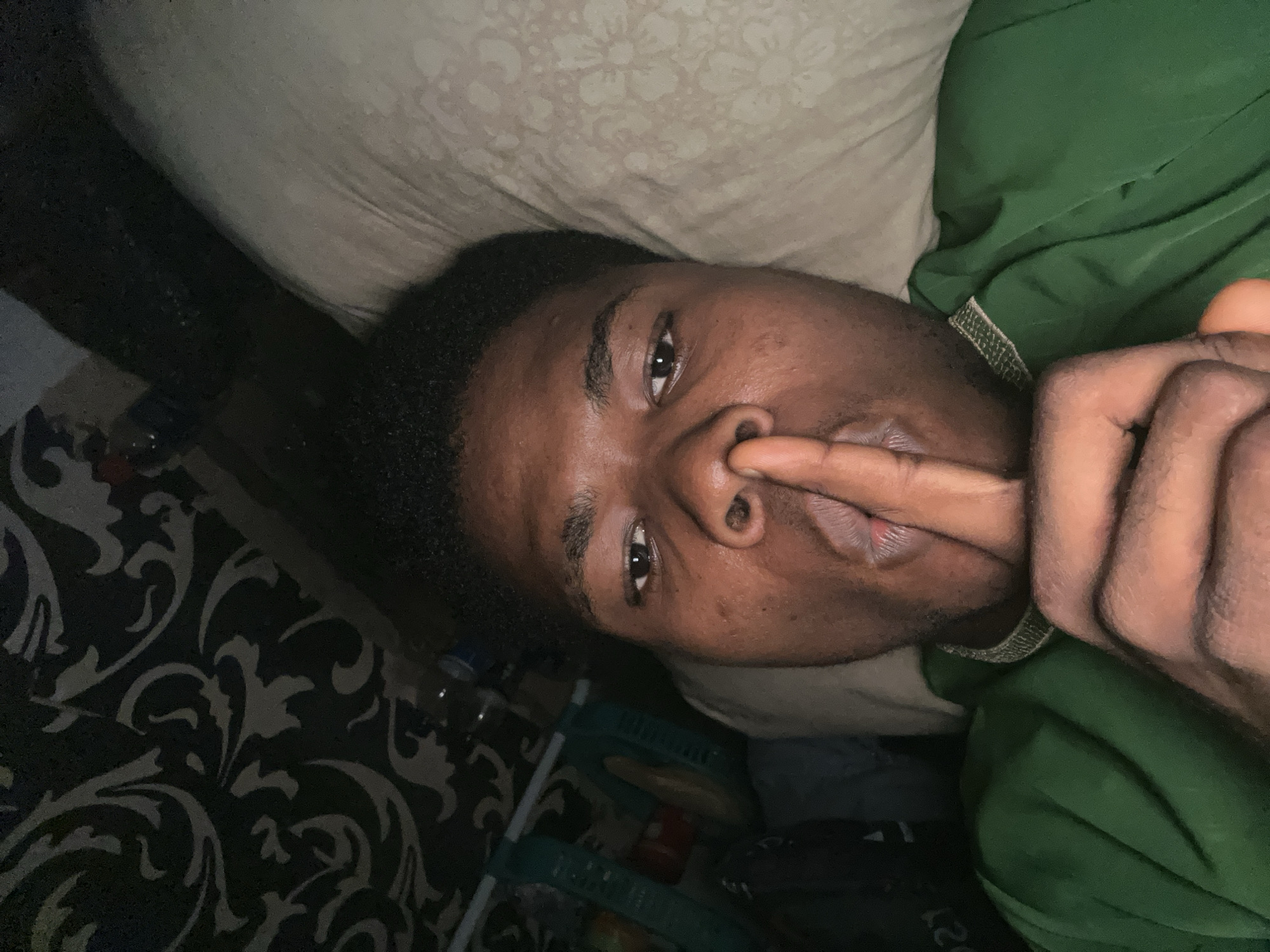 In Ihiagwa, Nigeria, a person with dark skin lies on their back on a light-colored, patterned pillow. They are wearing a green garment, possibly a hoodie or sweatshirt, which is draped over their left shoulder and chest. Their right hand is raised to their face, with a finger gently pressed against their lips, in a gesture that could indicate silence or contemplation. The person's head is slightly tilted, and their eyes are open, looking towards the camera. The background is dimly lit and features a dark, patterned wallpaper. To the left of the person's head, a green mesh basket filled with various items is visible, along with a few plastic bottles. The overall scene suggests an indoor setting, possibly a bedroom or living area.