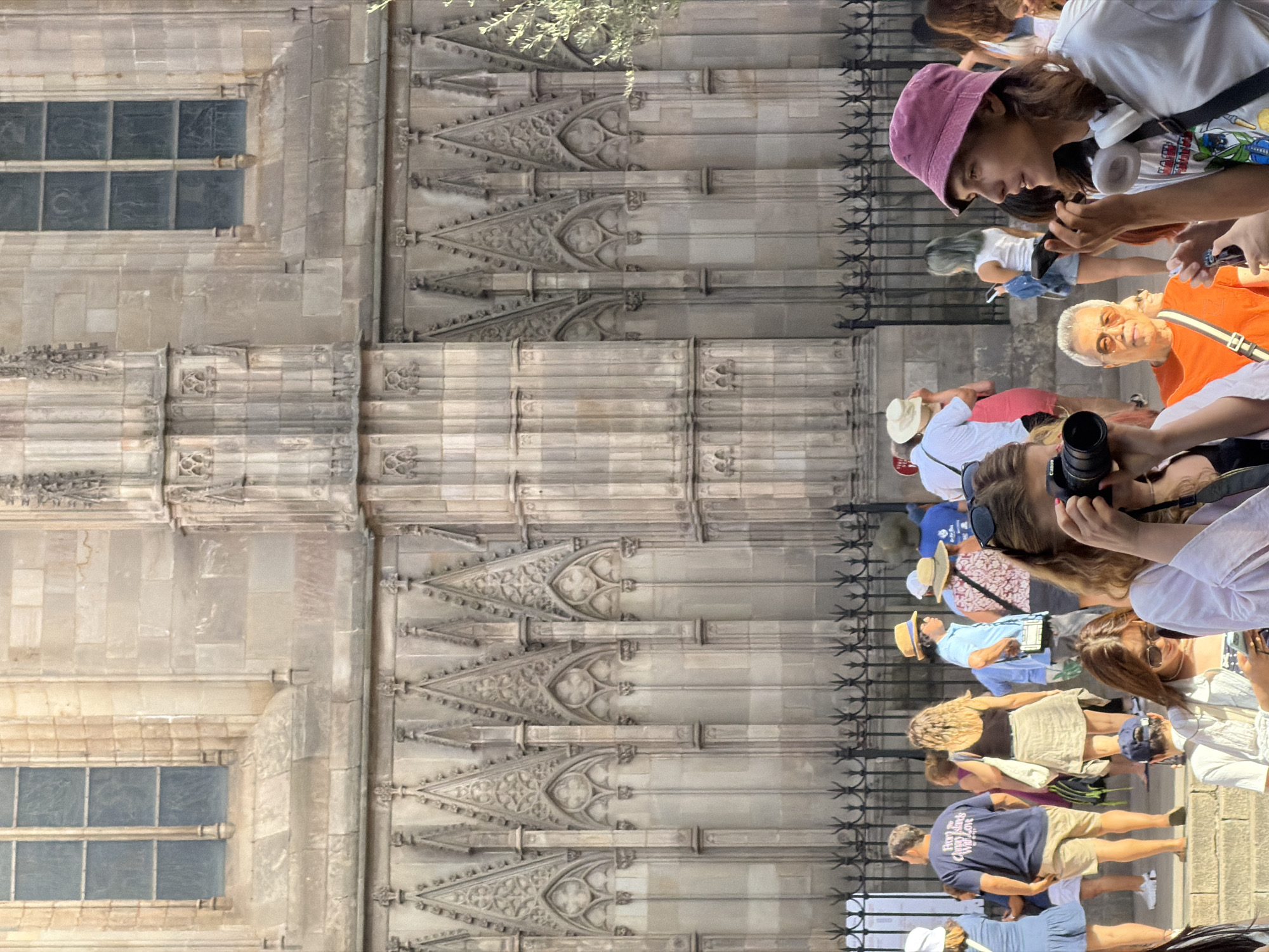 The image shows a group of people gathered in front of a large, ornate stone building, likely a cathedral, in Barcelona, Spain. The building's facade is characterized by intricate Gothic architectural details, including pointed arches, decorative carvings, and large windows with stained glass. A black wrought-iron fence runs along the base of the building.
In the foreground, a young person with long brown hair, wearing a pink bucket hat and a white t-shirt with graphics, is holding a smartphone to their ear and a camera in their other hand. They are looking to the right of the frame. To their left, another person is holding a black DSLR camera and looking down at it.
Behind them, a diverse crowd of tourists are milling about. Several individuals are wearing hats, including straw hats and a blue baseball cap. One person is wearing a navy blue t-shirt with white lettering that reads "FROM THE CANARY ISLANDS WITH LOVE". Another person is wearing a white top and a patterned skirt. A man in an orange t-shirt and sunglasses is visible. The people are in various poses, some looking at their phones, others talking, and some appear to be posing for photos.
The overall scene suggests a bustling tourist location, possibly during daylight hours, given the lighting and the presence of outdoor activities. The architecture strongly suggests a historical landmark, fitting with the prominent cathedrals found in Barcelona, such as the Barcelona Cathedral (Catedral de Barcelona) or the Santa Maria del Mar.